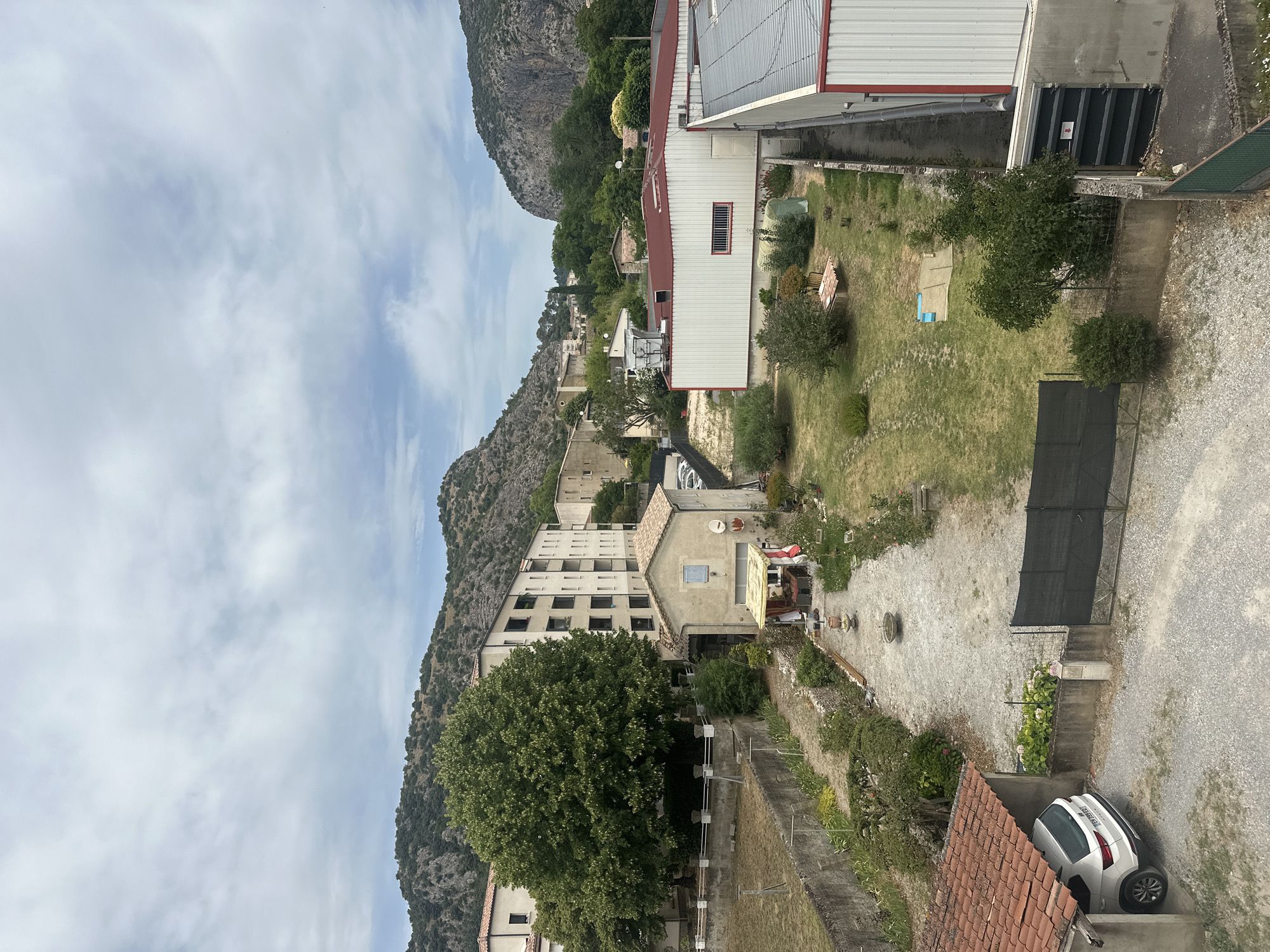 The image captures an aerial view of a residential and industrial area in Anduze, France. In the foreground, a white car is parked under a tiled roof carport. Adjacent to the carport is a gravel driveway leading to a black metal gate. Beyond the gate, a small garden with a lawn and several trees is visible. Further in the distance, a long, white industrial building with a red roof stands. Behind it, a cluster of residential buildings, some multi-story apartments and smaller houses, are nestled against a backdrop of lush green hills and rocky outcrops, characteristic of the Cévennes region. The sky is overcast with soft, diffused light. The scene depicts a typical semi-rural setting with a mix of residential and commercial structures.