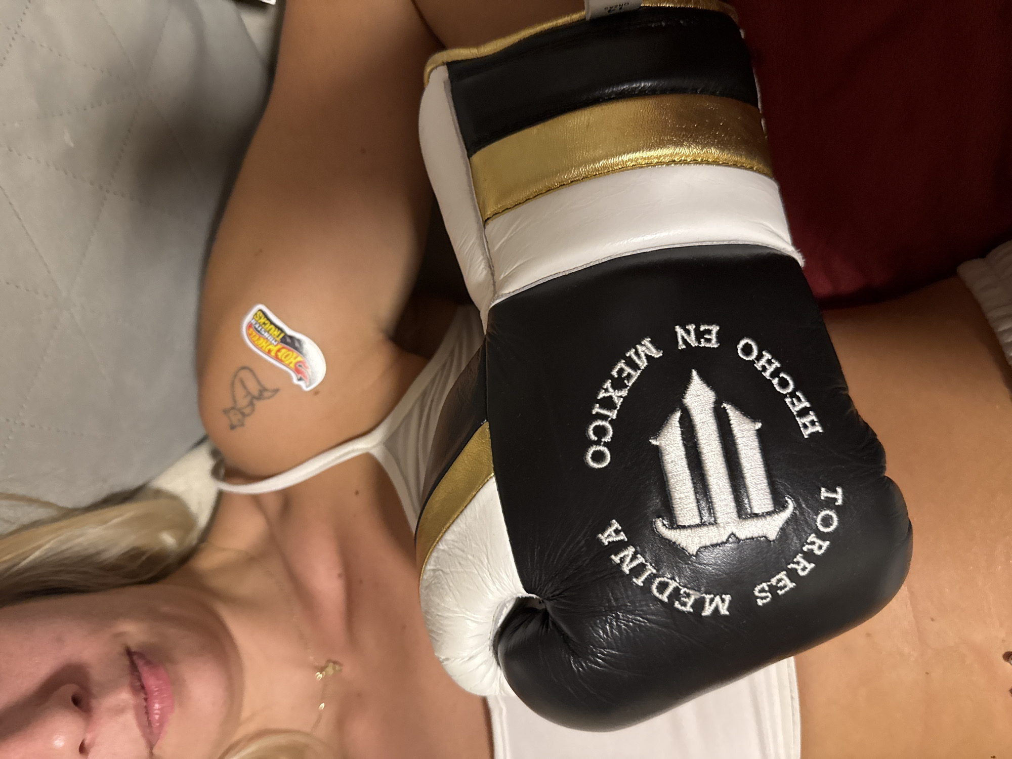 The image, captured in Plovdiv, Bulgaria, presents a close-up, top-down view of a woman holding a boxing glove. Her blonde hair is visible around her partially shown face, which includes her mouth and nose, revealing a slight smile. She is wearing a white tank top and a thin gold necklace with a small charm. On her upper arm, there is a prominent sticker featuring the words "HOT WHEELS MONSTER TRUCKS," alongside a small, simple tattoo.
The boxing glove, held or resting on her, is predominantly black with white accents and a metallic gold stripe. Embroidered on the glove in white text are the phrases "HECHO EN MÉXICO" (Made in Mexico) and "MEDINA TORRES," framing a distinct, stylized logo resembling a trident or an "M". The background includes a textured grey surface to the left and a solid maroon area to the right, suggesting an indoor setting and a relaxed pose. The lighting appears to be soft and diffused.