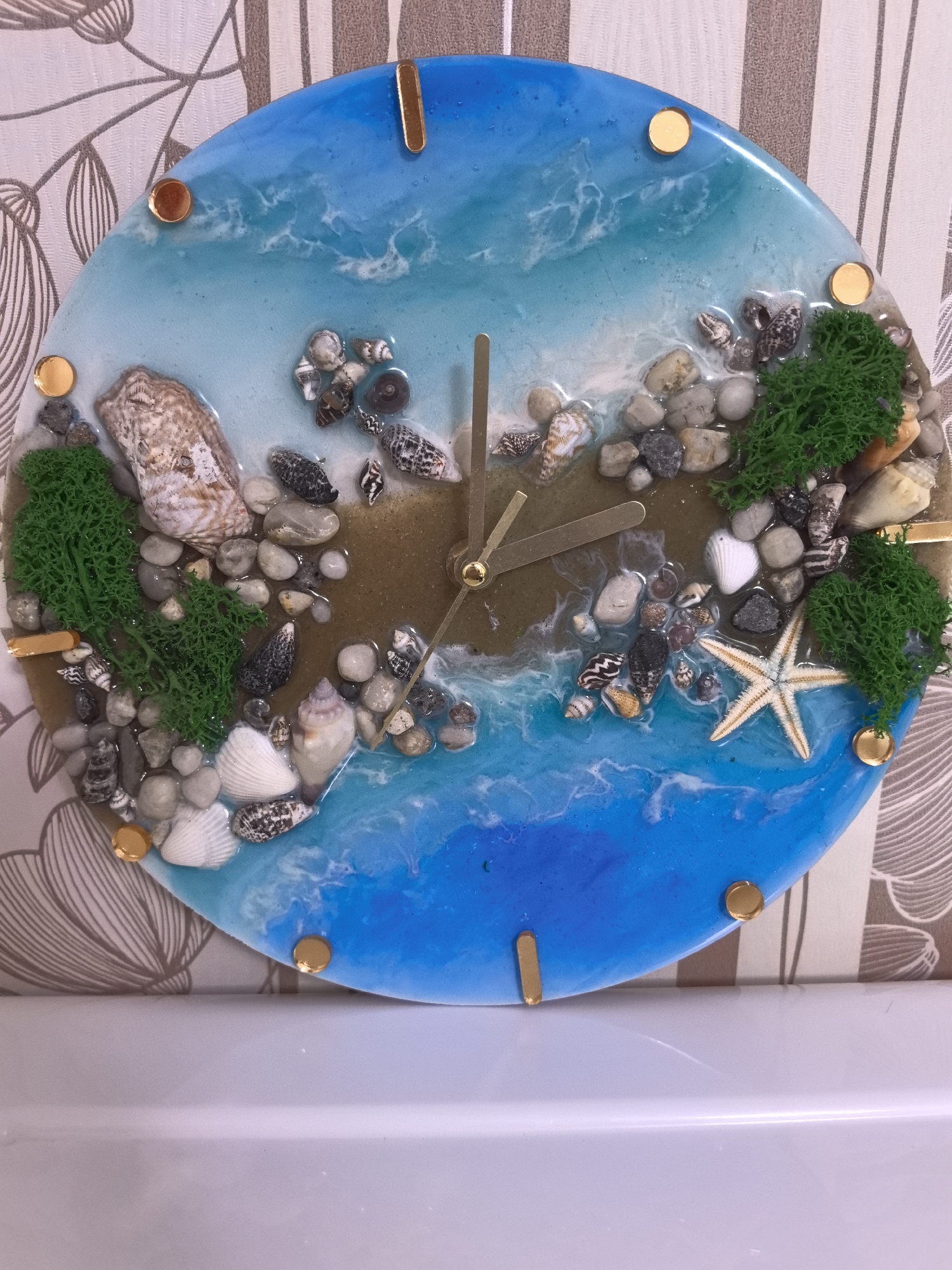 The image shows a round resin clock with a beach theme. The clock face is blue and brown, simulating water and sand. It is decorated with seashells, small rocks, green moss, and a starfish. Gold-colored round and rectangular markers indicate the hours. The clock hands are also gold-colored. The clock is placed on a white surface, likely a shelf or ledge, against a background of patterned wallpaper. The location, Парэчча, Belarus, is not discernible from the image itself.