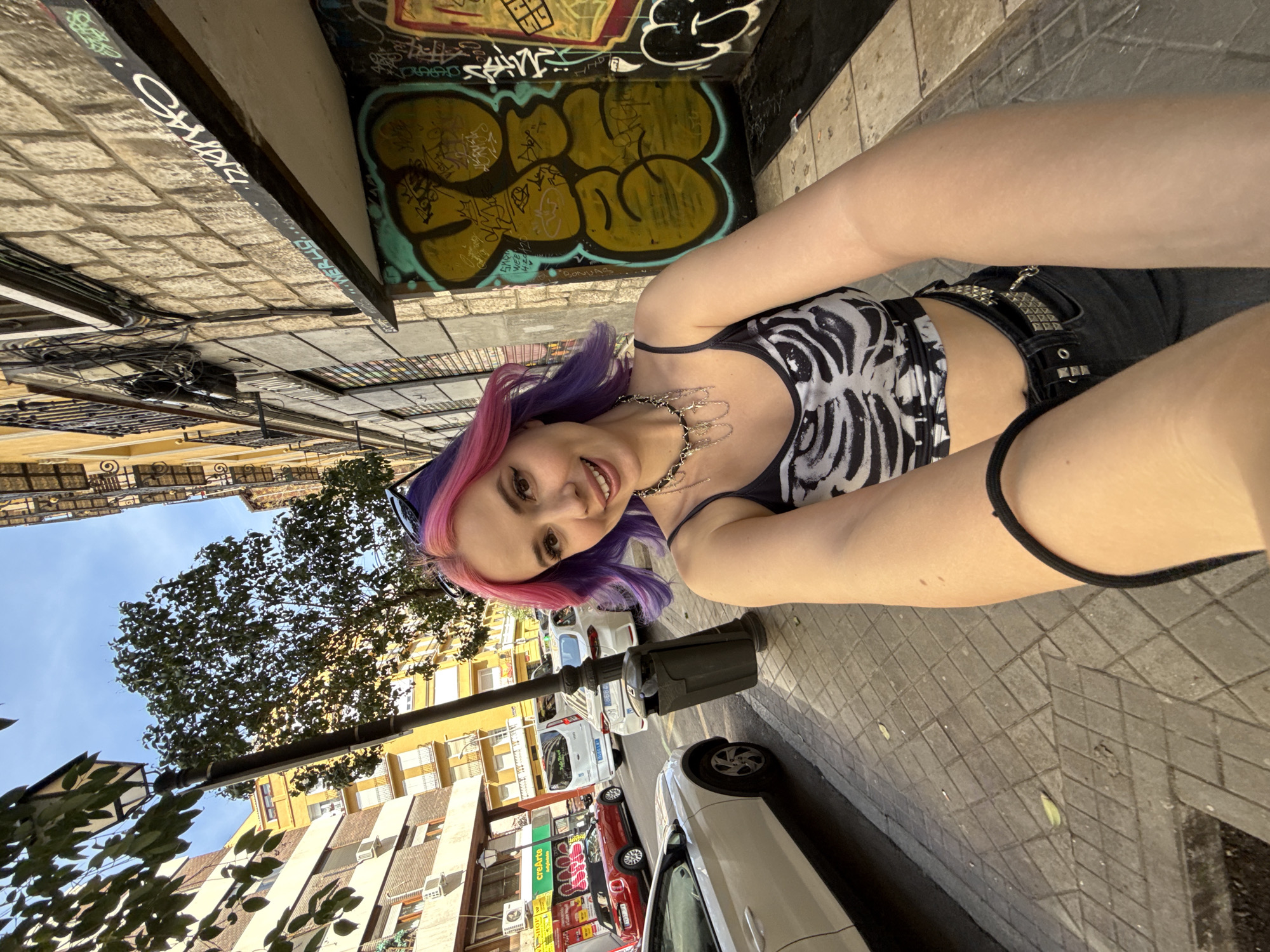 The image shows a woman with pink and purple hair wearing a black and white top, a studded belt, and black shorts, seemingly taking a selfie outdoors. The background indicates an urban setting, likely Madrid, Spain, suggested by the architectural style of the buildings and the street layout. Graffiti adorns a corner behind her, adding to the urban backdrop. Cars are parked along the street, and a tree's foliage is visible, contributing to the outdoor atmosphere.