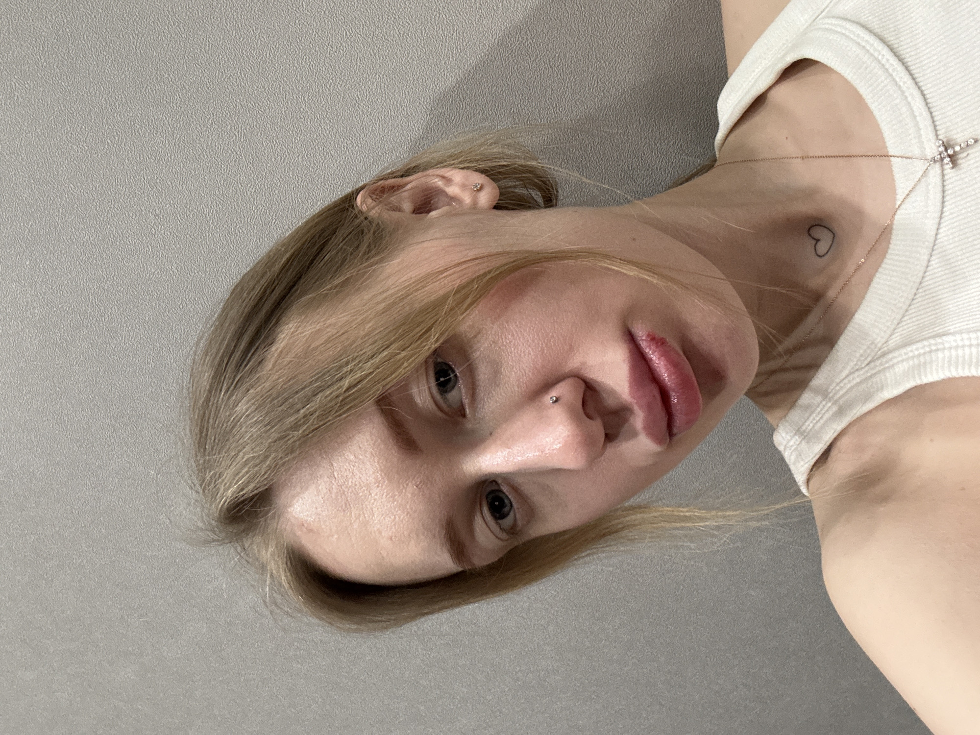 A close-up, head-and-shoulder-level indoor image depicts a fair-skinned female subject with blonde hair and blue eyes, looking towards the camera. The setting is an interior space with a plain, light grey textured wall serving as the background. The subject's left nostril features a small silver stud piercing. Her lower lip exhibits a visible lesion or sore with associated redness on its left side. A small black outline heart tattoo is present on her neck, slightly to the left of the centerline. She wears a thin, gold-colored chain necklace with a small, jewel-encrusted cross pendant resting on her upper chest, over a white ribbed tank top. A small stud earring is visible in her left ear. The location is specified as Rostov-on-Don, Russia.