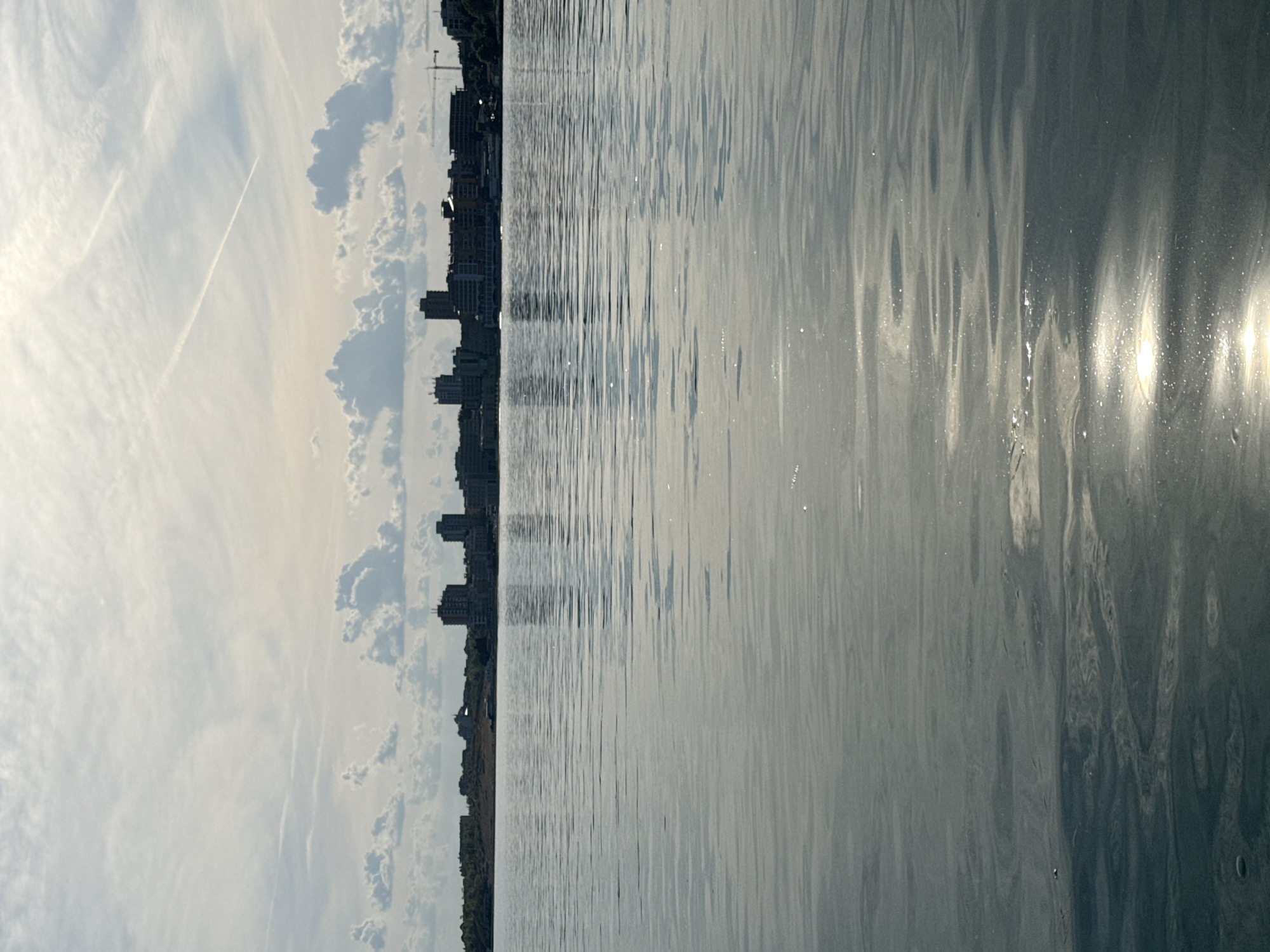 The image shows a waterfront view in Nesebar, Bulgaria. The water is calm, reflecting sunlight. A line of buildings, including several tall apartment blocks and a construction crane, defines the horizon. The sky is partially cloudy with contrails visible. The image is oriented sideways.