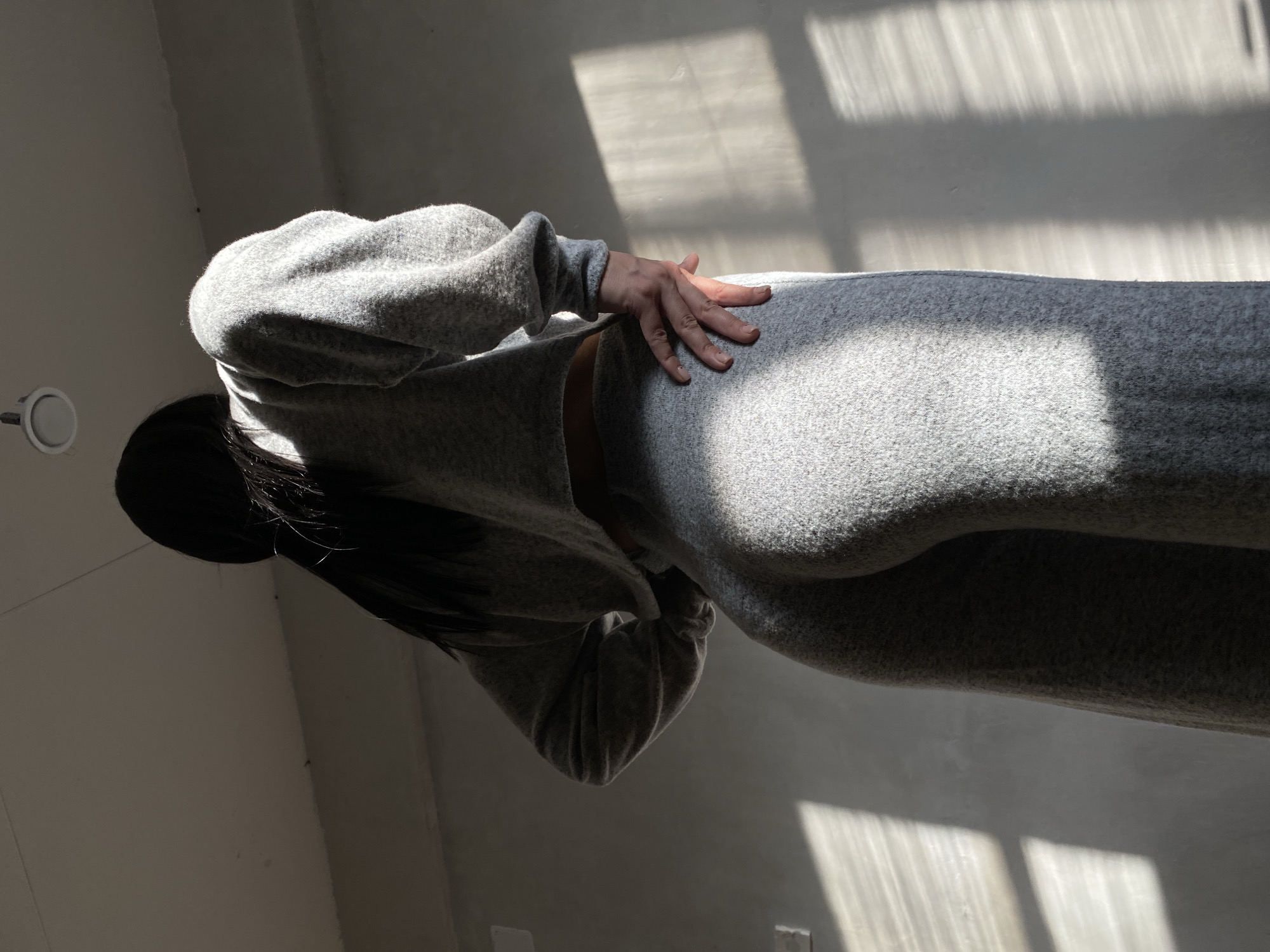 A person with long black hair wearing a gray sweatsuit is indoors. The location is unknown, and there is no information to suggest it is in Votkinsk, Russia. Light streams in from a window, casting shadows on the floor and wall. The person's hand rests on their lower back/buttocks area. A light fixture is visible on the ceiling.