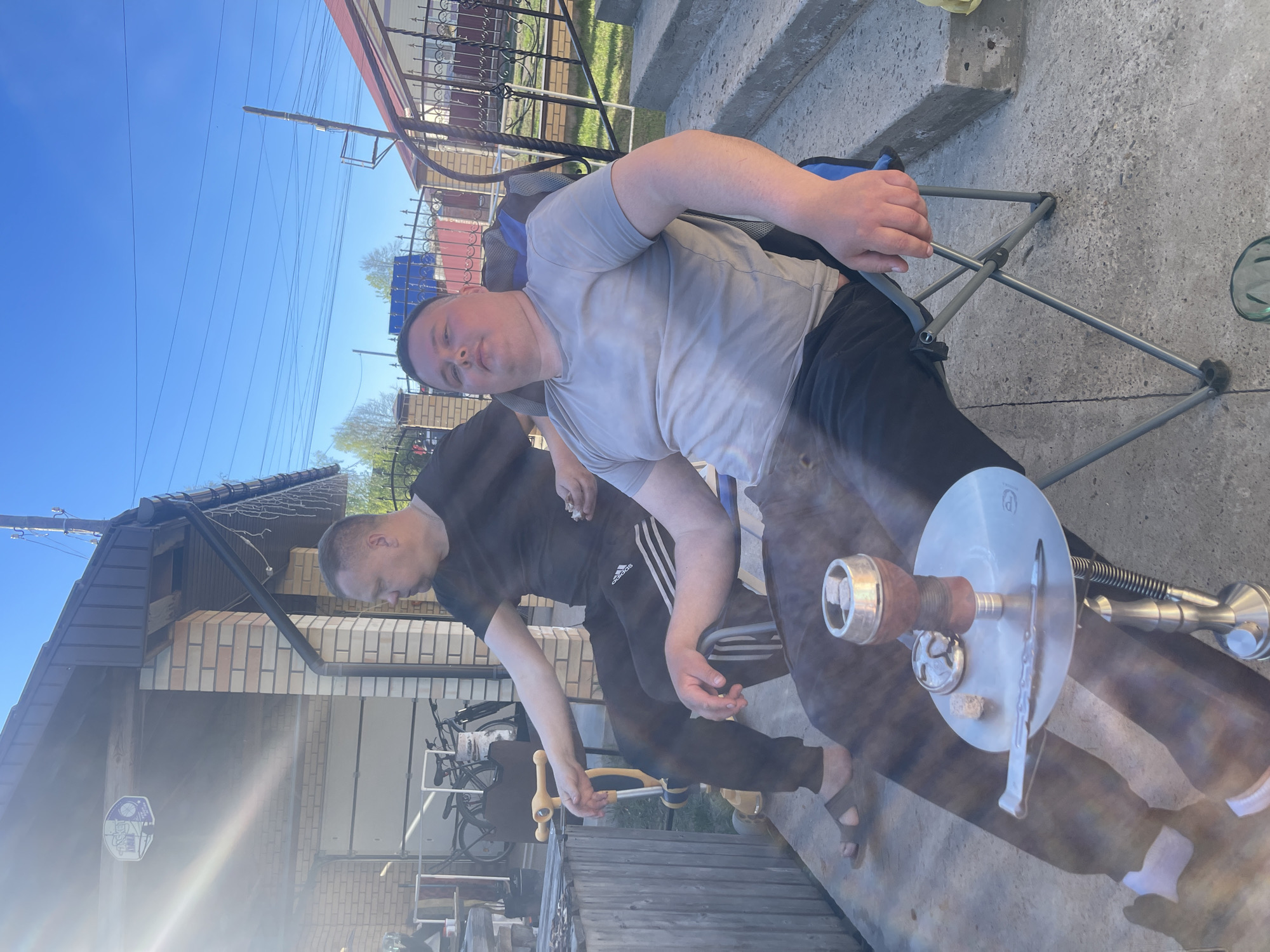 Two men are outdoors, likely in a residential area. One man is seated in a blue and grey camping chair wearing a light grey t-shirt and black pants. The other man is standing, wearing a black t-shirt with white stripes down the side and black track pants. He is holding a yellow object in his right hand. In front of them is a hookah. The setting appears to be the city of Chaikovsky, Russia, based on the architectural style of the brick house in the background. There is a house behind the men and a fence near them.