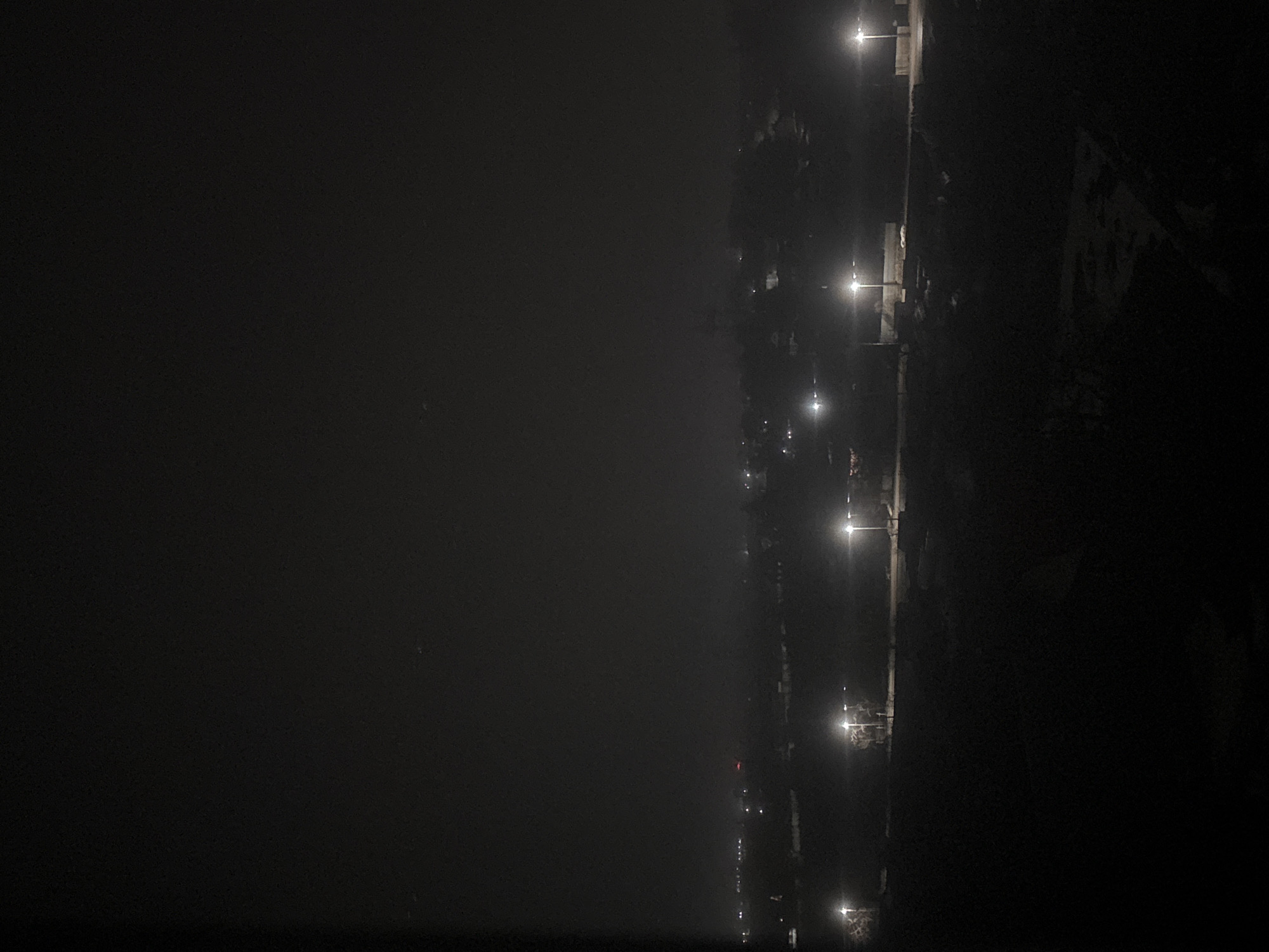 At night in Chaikovsky, Russia, streetlights illuminate a roadway. Fog obscures the view to the left. Houses and foliage appear on the right, shrouded in darkness.