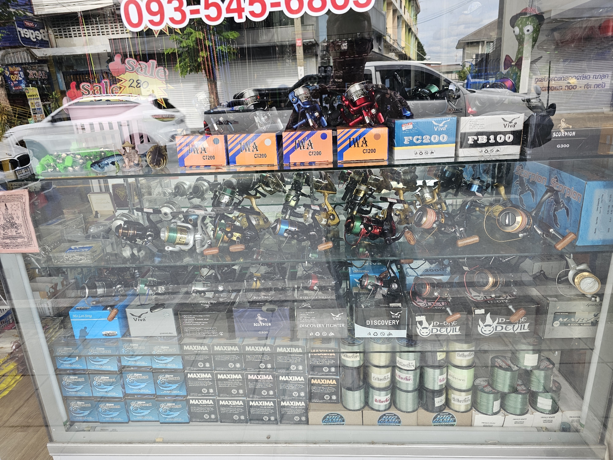 The image shows a display window of a fishing tackle shop in Bangkok, Thailand. The window is filled with various fishing reels, fishing lines, and other fishing equipment. Some of the products are packaged in boxes with Thai and English text, and some feature graphics of fish or other aquatic life. The shop also appears to be selling discount items, as indicated by a large red "Sale" sign visible through the window. In the background, reflections in the glass show buildings and a street scene, typical of an urban environment in Bangkok. There are no people clearly visible in the image, but the display suggests a retail setting catering to fishing enthusiasts.