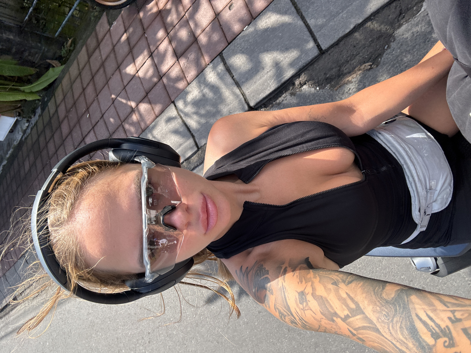 A high-angle, outdoor shot depicts a woman, presumably in North Kuta, Indonesia, wearing black headphones and transparent sunglasses. She has blonde hair and a visible tattoo on her right arm. She wears a black zip-up top and a light-colored waist bag. The background includes a sidewalk and a portion of a street. The pavement material is concrete and brick. The lighting suggests daytime conditions.