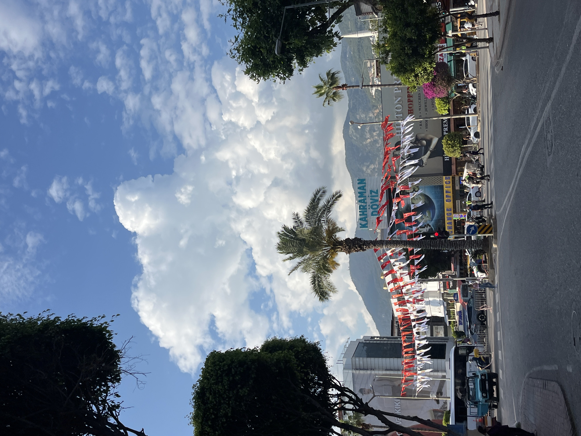 The image shows a street scene in Alanya, Turkey. The view is from a low angle, looking up towards a street lined with buildings, palm trees, and decorations. A mountain is visible in the background, partially obscured by clouds. Turkish flags are strung across the street. A large advertisement is on the side of a building for "KAHRAMAN DOVIZ". Some shops and vehicles are visible on the right side of the street. The sky is blue with large, fluffy clouds. Trees frame the foreground of the image. The scene appears to be taken on a sunny day.
