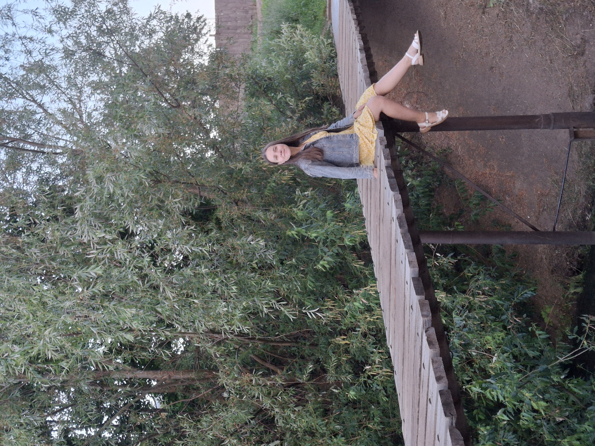 A young woman with long brown hair sits on the railing of a wooden bridge, her legs dangling over the edge. She wears a denim jacket over a yellow floral dress and white sandals. The bridge appears to be made of weathered wood planks. She looks towards the camera with a slight smile.
The bridge is surrounded by lush green trees and foliage, suggesting an outdoor, natural setting. The lighting indicates it is daytime, possibly late afternoon given the soft light. The scene has a casual and relaxed atmosphere. The background shows more greenery and a hint of a wooden fence. The location is identified as Chaikovsky, Russia. There is no visible text in the image.