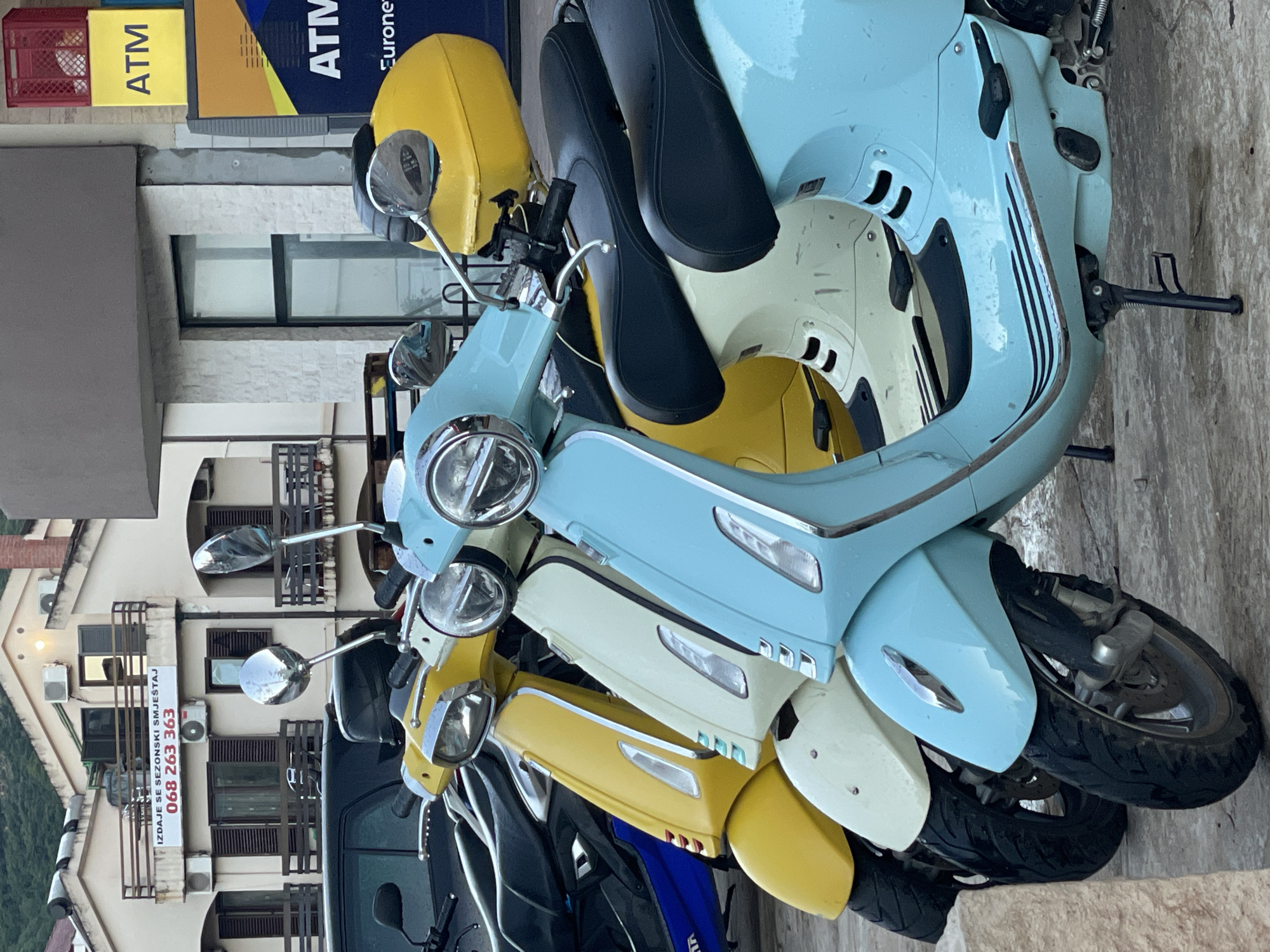 A row of parked Vespa scooters in various colors (blue, yellow, and cream) is captured in an outdoor setting. An ATM sign can be seen on a building in the background. The context suggests a location in the city of Unknown, Unknown.