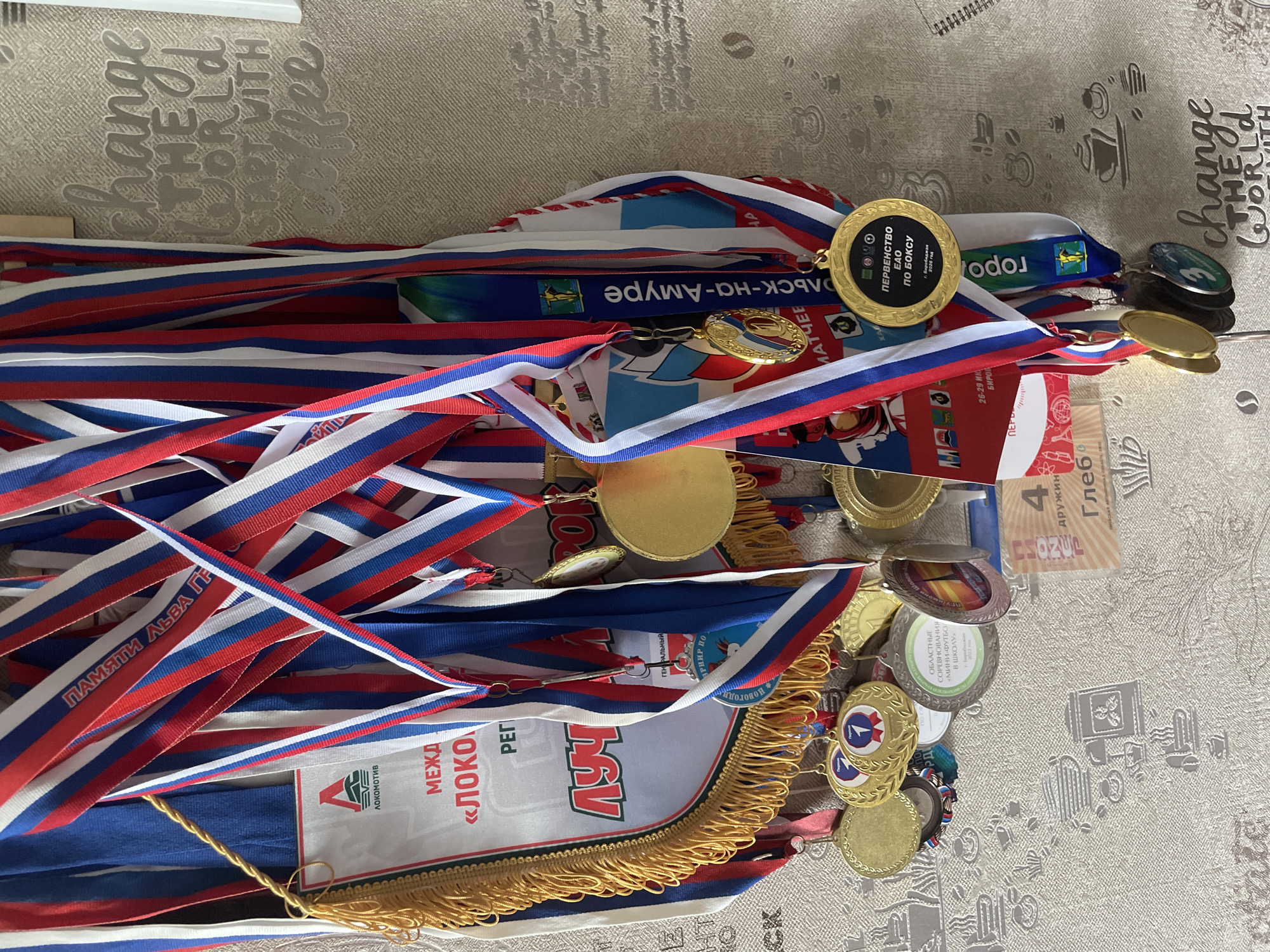 The image shows a collection of medals and awards hanging on a wall with textured wallpaper. The medals have ribbons in the Russian flag colors of white, blue, and red. Some of the medals are gold, silver, and bronze colored. One prominent medal has Russian text that translates to "Championship of the Far Eastern Federal District in Boxing". Another medal has text that says "Regional Sports Competition in Mini-Football in School". A large banner or flag with gold fringe is also visible, with Russian text that includes "LOCOMOTIVE". There are also some decorative elements on the wallpaper, including a phrase that reads "change THE WORLD START WITH coffee" and drawings of coffee cups. The overall scene suggests a display of athletic achievements.