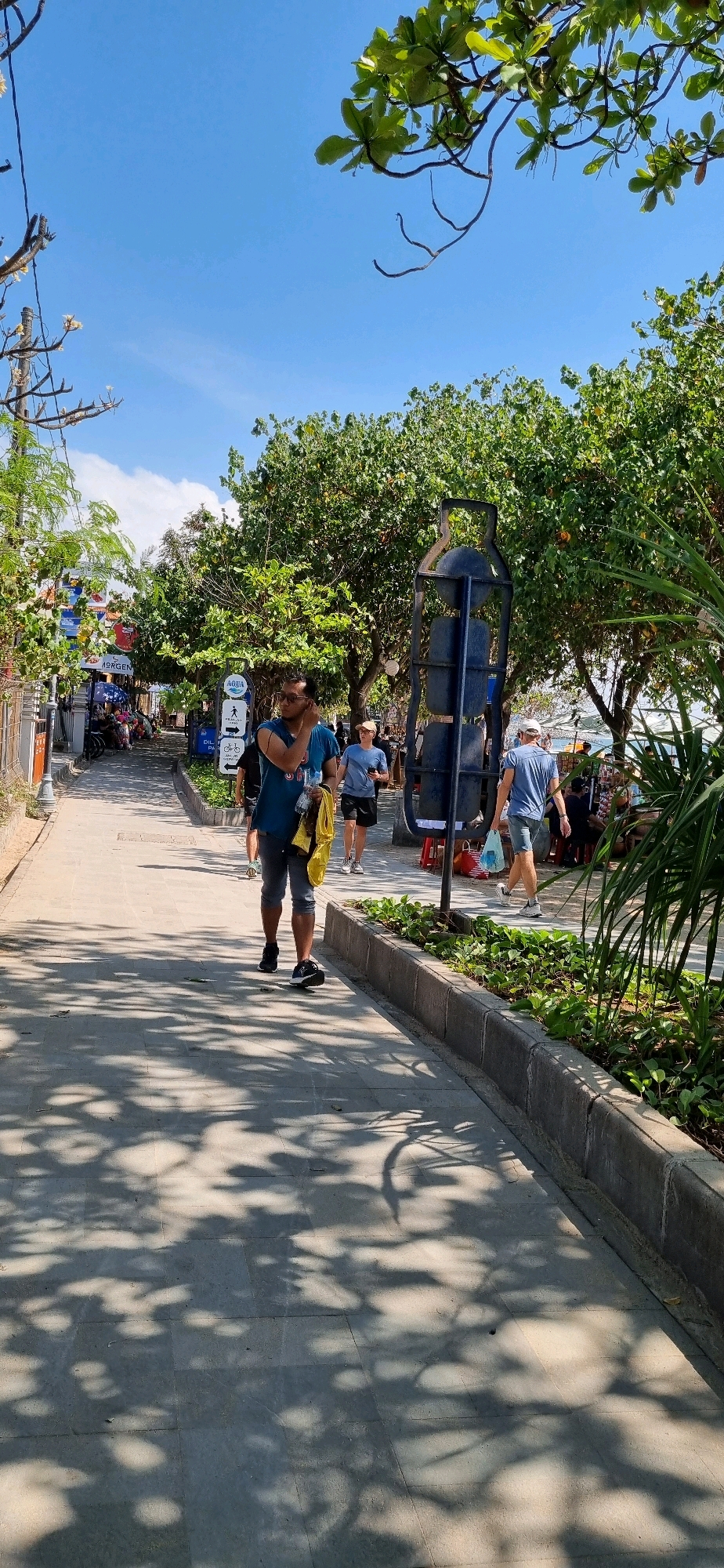 This is a vertical, eye-level, full-shot photograph of a paved walkway in Denpasar, Indonesia, likely a promenade near a beach or tourist area, given the presence of palm trees, lush foliage, and what appears to be a light ocean breeze. The sun is bright, casting dappled shadows from overhead trees onto the walkway.
In the foreground, a man wearing a blue t-shirt and grey shorts is walking towards the camera, holding a yellow bag in his right hand and an object to his ear with his left. He is wearing black sneakers. Behind him, also walking away from the camera, is a person in a blue t-shirt and black shorts, wearing a light-colored cap and white sneakers. Further back, another person in a blue t-shirt and light-colored shorts is walking away from the camera, carrying a plastic bag in their right hand.
To the left of the walkway, there are buildings with shops, awnings, and signs, indicating a commercial area. To the right, a raised garden bed with green plants lines the walkway, and beyond that, more trees and foliage can be seen. A dark, tall, bottle-shaped metal structure with a spherical top is positioned in the middle ground on the right side of the walkway. In the background, there are glimpses of more trees, a body of water, and possibly more distant structures. The sky is clear and blue. The overall atmosphere is one of a sunny day in a tropical urban environment.