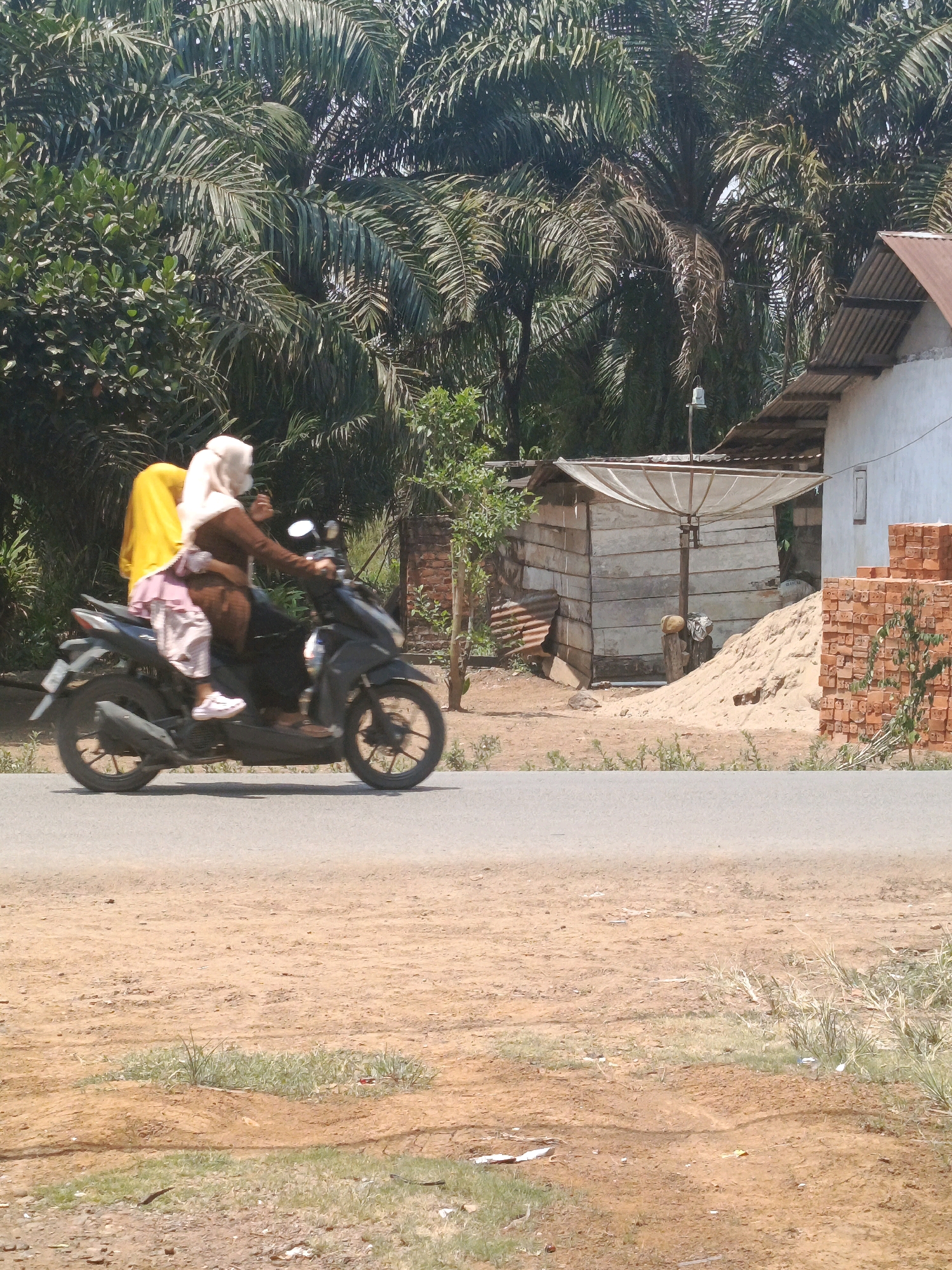 In Megang Sakti I, Indonesia, a woman wearing a light-colored hijab and a brown tunic rides a dark-colored motorcycle. She is carrying a child on her lap who is wearing a pink and white striped outfit and a yellow hijab. The motorcycle is moving on an asphalt road. In the background, there are palm trees, a small wooden building with a satellite dish, a pile of sand, a stack of bricks, and a white building with a metal roof. The ground on the side of the road is unpaved and dusty, with some sparse vegetation.