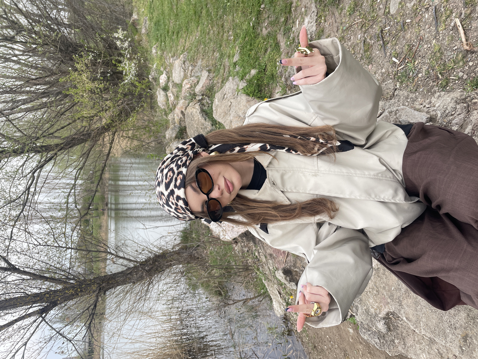 A woman stands on a rocky embankment, near a body of water lined with leafless trees. She wears a light tan jacket, brown pants, sunglasses, and a leopard-print headscarf. She is making a rude gesture with both hands, each adorned with a large gold ring.
While the exact location is difficult to determine without further information, the scene may be in or near a park in Sofia, Bulgaria, given the combination of water, trees, and rocky ground.