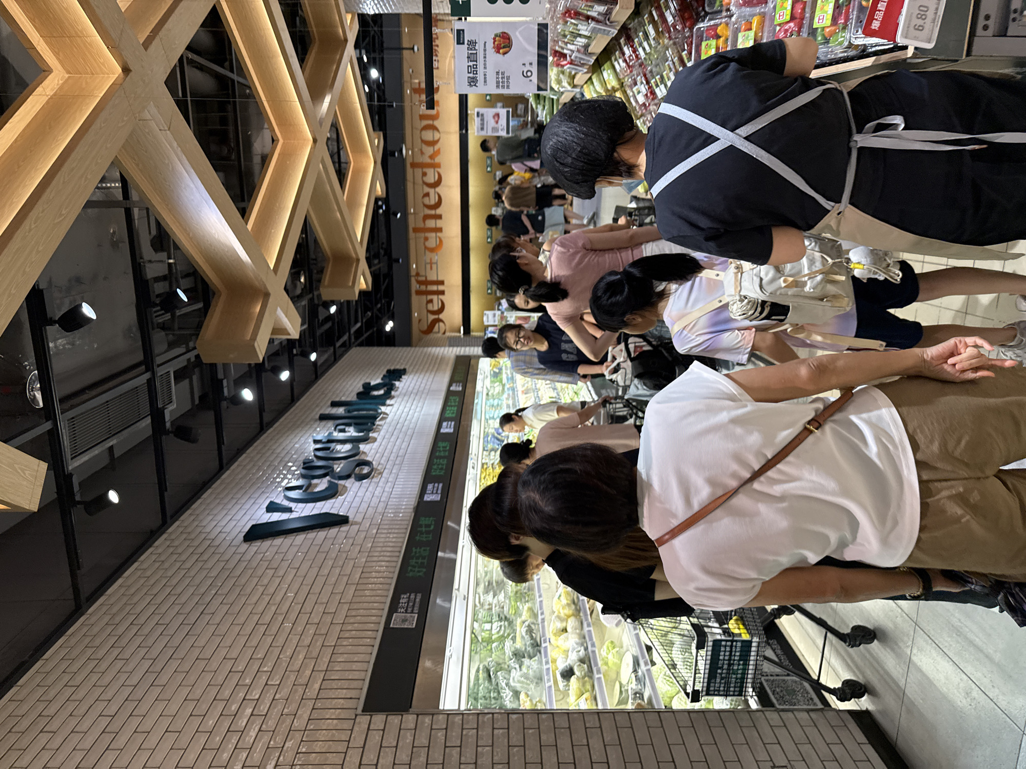 The image depicts a grocery store aisle in Haizhu District, China, with a queue of people waiting at a self-checkout area. In the foreground, a person wearing a white t-shirt and brown shorts stands with their back to the camera, a tan crossbody bag slung over their shoulder. To their left, another person in a black t-shirt is partially visible. A younger person with a backpack adorned with a grey plush toy stands further ahead in the queue. In the background, various shoppers are seen, some with shopping carts, and aisles of produce and other groceries are displayed. Above the checkout area, a sign reads "Self-checkout" in large orange lettering, and a brick wall with the word "Vegetables" in large, stylized letters is visible to the left. The lighting suggests an indoor environment, possibly a modern supermarket.