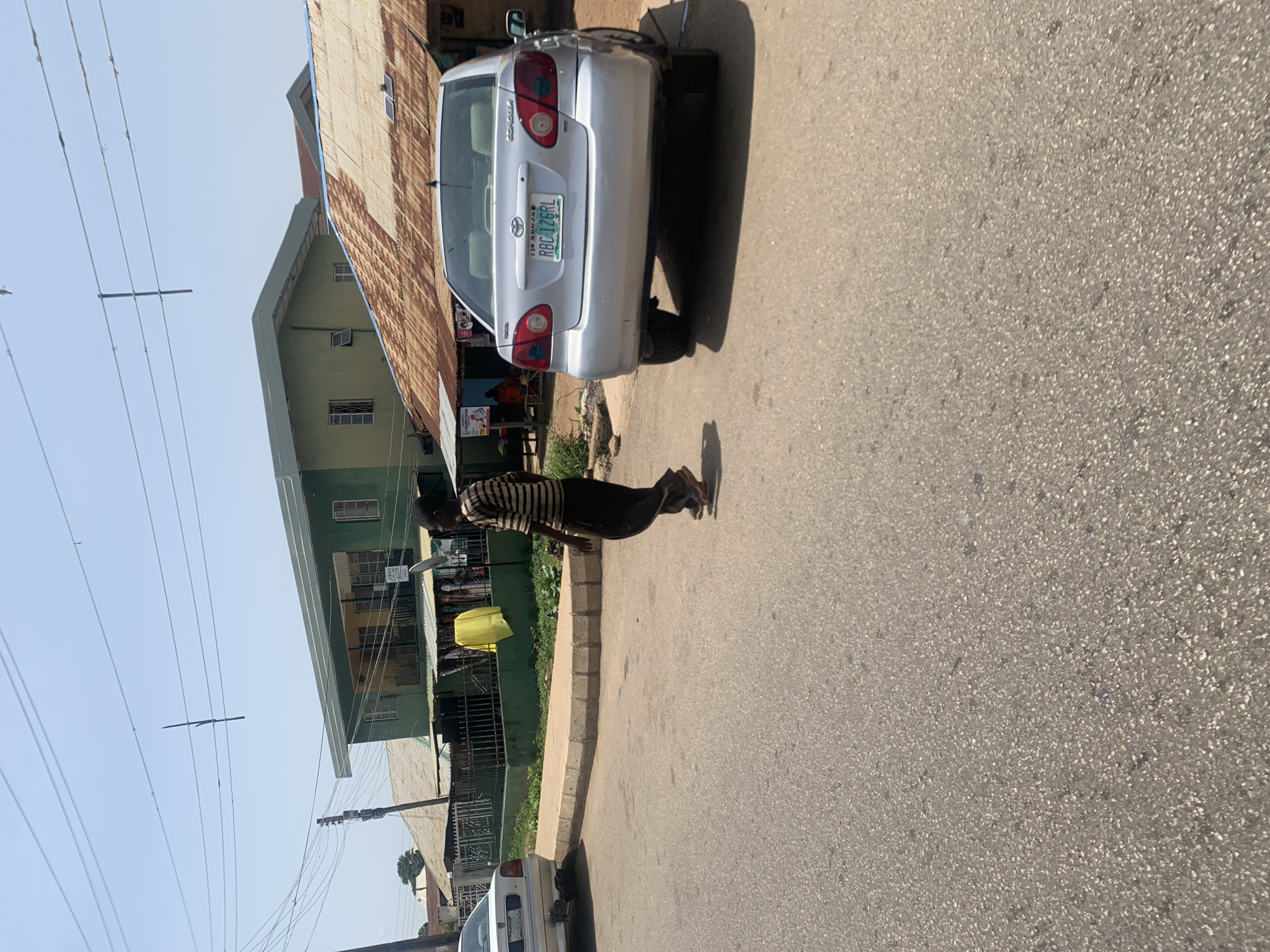 A man in a striped shirt and dark pants walks on a paved street in front of a silver Toyota Corolla with a Nigerian license plate, RBC-126RL. The car is parked on the side of the road, and a sidewalk with a curb is visible behind it. In the background, there are buildings with green and rusty corrugated metal roofs, some with awnings and open windows. Clothes are displayed on racks in front of one of the buildings. Power lines crisscross the sky above the street and buildings. The scene appears to be in an urban area, possibly a marketplace or a residential street, in Jos, Nigeria.