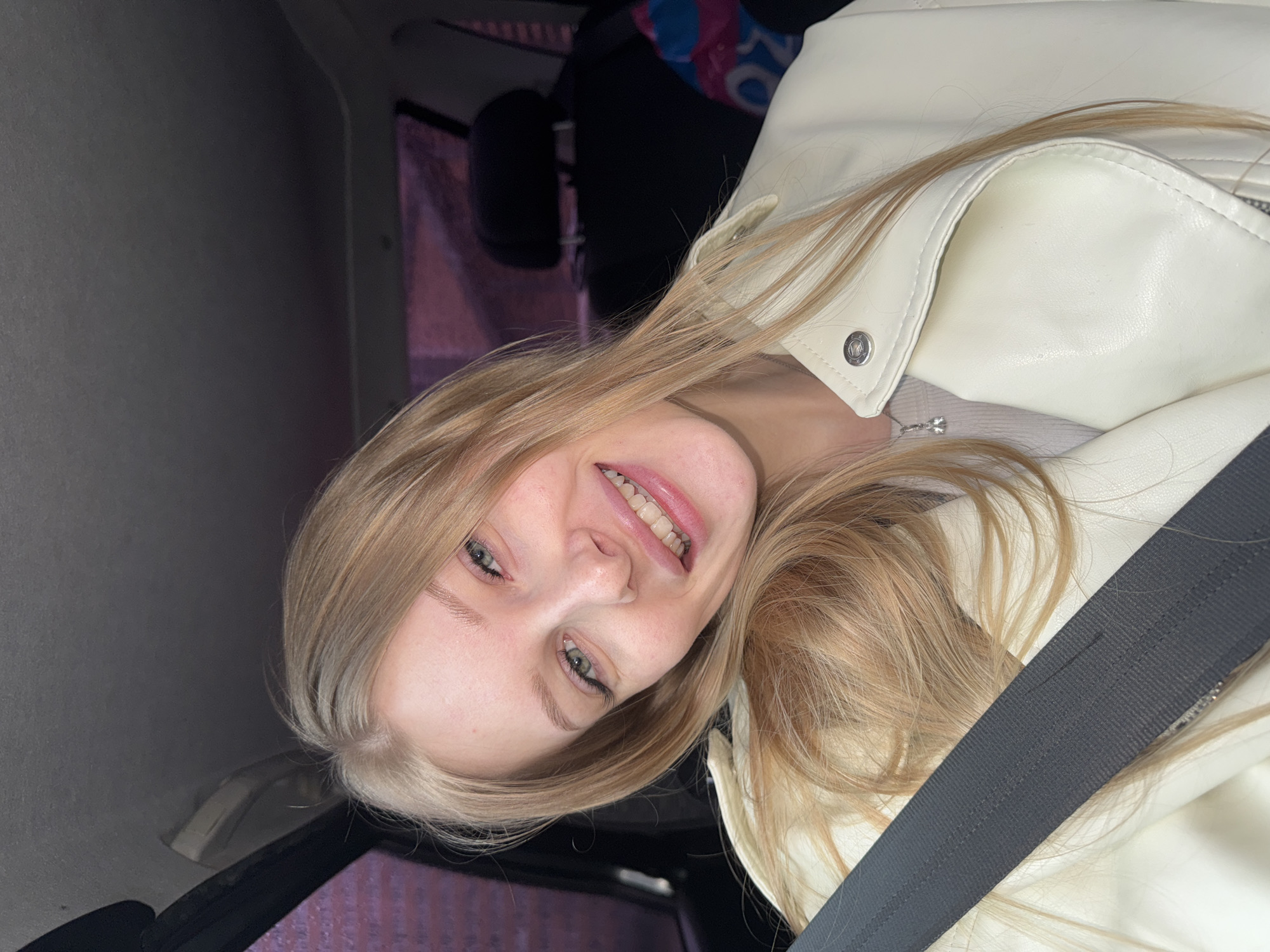 A young, fair-skinned woman with blonde hair is smiling at the camera while wearing a seatbelt and a light-colored jacket in what appears to be a car. The specific location in the city of Perm, Russia, cannot be determined from the image.