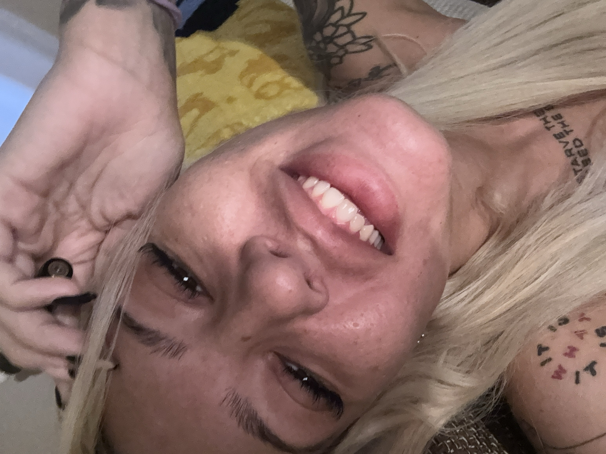 A close-up portrait shows a person with blonde hair, fair skin, and multiple tattoos, lying down and smiling. The image is taken from an elevated, angled perspective, with the subject's head tilted to the right. The person's face is central, displaying a wide smile with visible teeth, and bright, open eyes. Long, light blonde hair frames the face and cascades around the head.
Notable details include several tattoos:
- On the neck/collarbone area, script reads "STARVE THE / FEED THE".
- On the right upper arm/shoulder, a circular tattoo contains abstract shapes and letters, including "M I T", with other indecipherable symbols around them. Further up the arm, another tattoo depicts a floral or abstract design.
One hand is visible in the upper left corner, with fingers wearing black nail polish, holding a small, dark, cylindrical object, possibly a vape pen or a makeup tool. The background suggests an indoor setting, likely a bed or sofa, with glimpses of a yellow patterned fabric (possibly a pillow or blanket). The person appears relaxed and happy, conveying an emotion of contentment. The lighting is soft and ambient. The setting is in Plovdiv, Bulgaria.