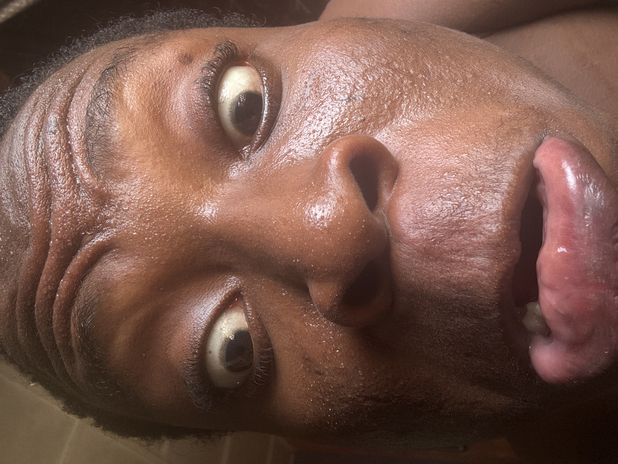 A close-up, low-angle, medium shot depicts the face of a Black male individual. The person's eyes are wide open, looking upwards. The mouth is also open, with the tongue protruding. The skin appears moist, possibly due to sweat. The location is Unknown, Unknown.