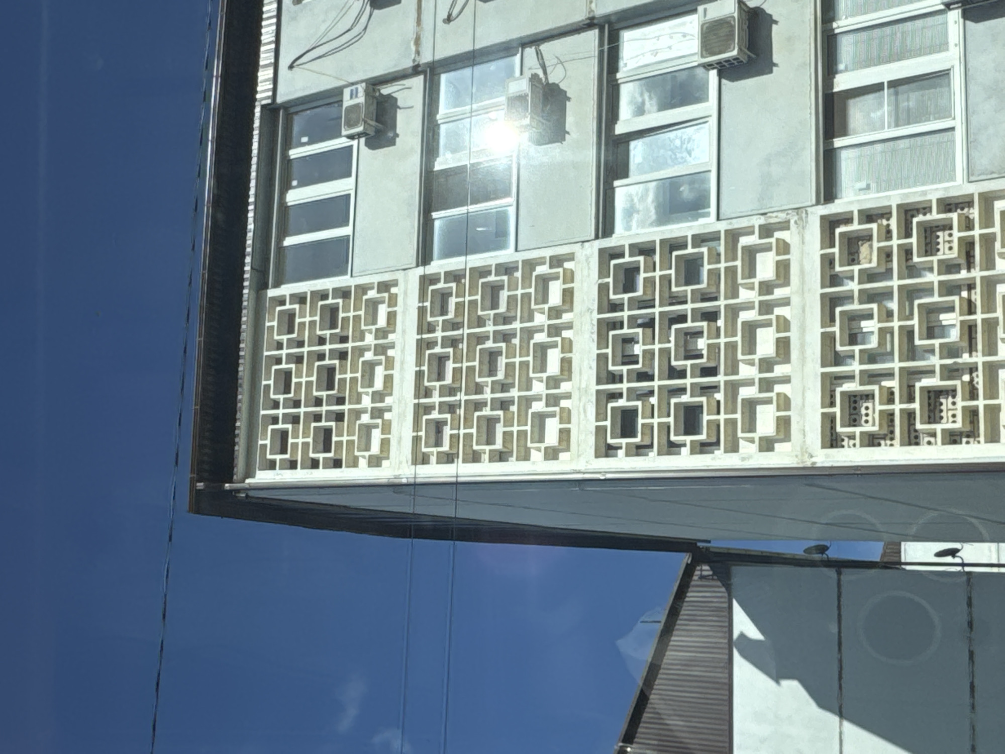The media file captures a close-up view of a building facade, likely in an urban setting. The building features a distinctive decorative screen composed of repeating geometric square patterns, made of a light-colored material. Above this screen, there are windows with multiple panes, some of which appear to be open. Above the windows, there are air conditioning units mounted on the exterior wall. Wires are also visible running along the top of the building.
The lower part of the image shows a reflective surface, possibly another building or glass, mirroring the sky. The sky is a clear, deep blue, suggesting a sunny day. The lighting indicates it's daytime, with sunlight reflecting off the building's surfaces and the windows. The angle of the shot, looking upwards at the building, and the reflective surfaces suggest the photo might have been taken from a lower vantage point, possibly from across a street or from a different building. The architectural style, particularly the decorative screen, might offer clues to the era or region the building is from. No people or specific activity are evident in the frame. The overall impression is one of architectural detail and the interaction of the building with its environment.