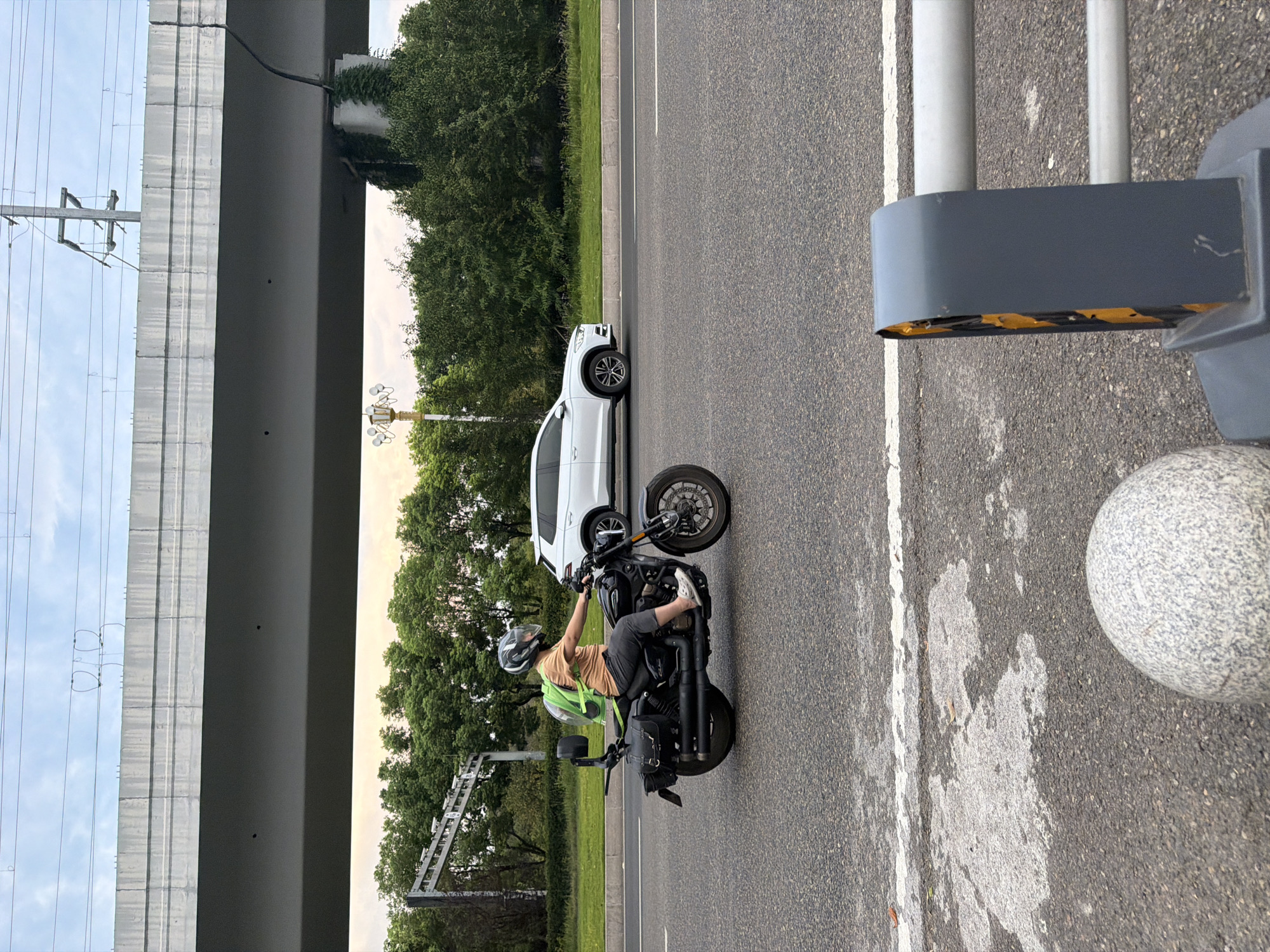 In Jiangjin, China, a person wearing a helmet and a light orange shirt rides a black motorcycle on a paved road. The motorcycle has saddlebags on its sides and a clear backpack attached to the rear. The person is also wearing dark pants and white slip-on shoes. To the right of the motorcycle, a white SUV is visible. Above the road, a large concrete overpass runs horizontally across the frame, supported by columns. Green trees line the roadside behind the motorcycle and the SUV. In the foreground, a grey spherical bollard and part of a grey barrier are visible. The sky above is cloudy.