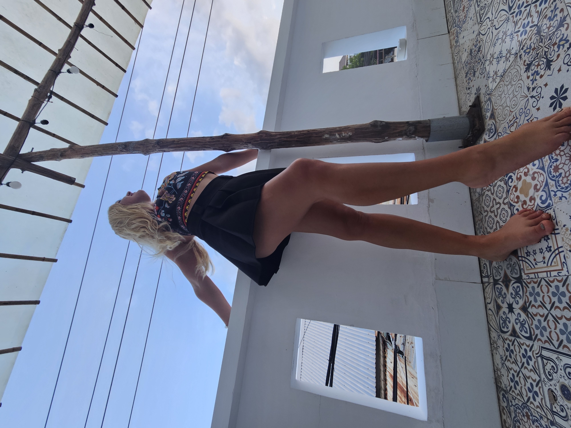 A low-angle, full-body shot shows a young Caucasian woman standing on a tiled patio in Phu Thuy Ward, Vietnam. The woman has long, blonde, wavy hair and is wearing a colorful, patterned crop top and a black pleated skirt. Her arms are extended outward as she leans against a white wall, and she is looking up at the sky. The patio has a detailed, colorful tile floor. Behind her, a rough wooden pole supports a canopy made of white fabric and bamboo. Two square openings in the white wall reveal a view of corrugated metal roofs and some distant buildings. The sky is a light blue with scattered clouds.