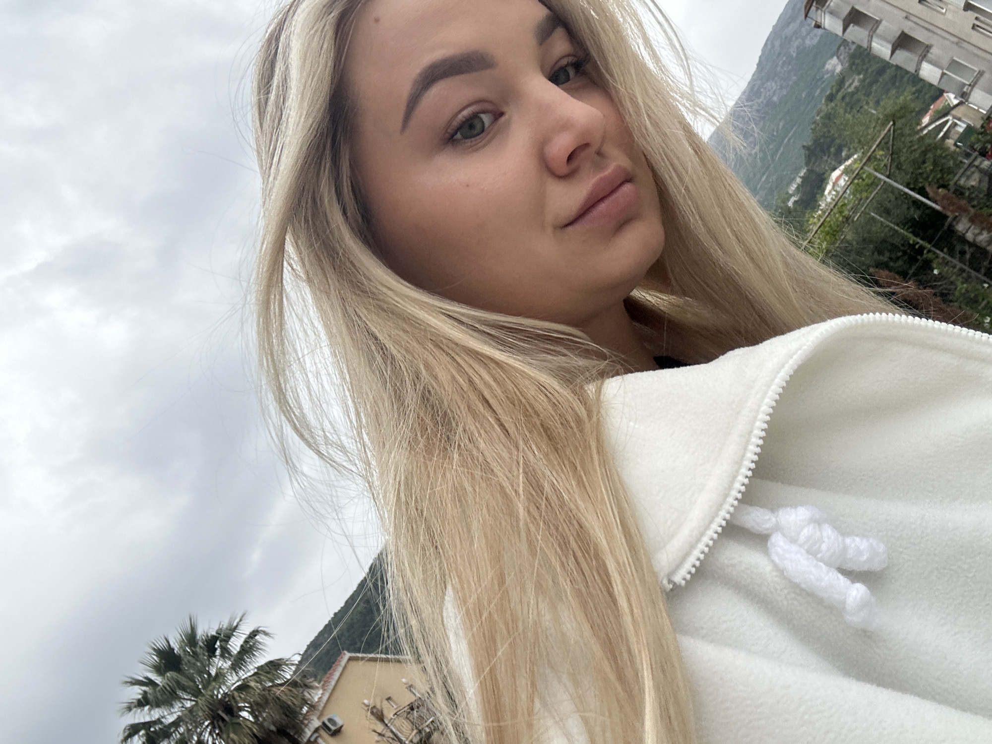 A young woman is featured in a close-up, eye-level selfie or portrait, looking directly at the camera with a slight pout. She has long, straight blonde hair that frames her face, fair skin, and green eyes, with well-defined eyebrows. She is wearing a white fleece jacket with a zipper and visible white drawstrings.
The setting is outdoors during the daytime under an overcast sky, suggesting cloudy weather. In the background, a large mountain range rises, covered in lush green vegetation. To the right, parts of a multi-story building with balconies are visible, typical of coastal architecture. Below and to the left, a large palm tree stands prominently against the cloudy sky, and another building with a visible air conditioning unit can be seen. The overall impression is a scenic, possibly coastal, location consistent with Budva, Montenegro. There is no visible text in the image.