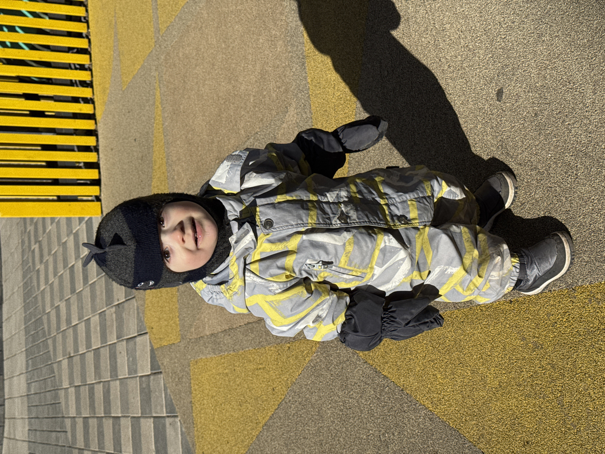 A small child wearing a gray and yellow snowsuit, black mittens, and a black hat with a fin-like decoration, is lying on a paved surface with geometric designs in yellow and gray tones. The child is looking directly at the camera. To the left of the child is a section of a yellow metal bench. The child's shadow is cast to the right, suggesting the light source is from the left. Based on publicly available information, it's plausible this photo was taken in a playground or recreational area in Izhevsk, Russia, given the likely clothing worn by the child is appropriate for a cold climate.