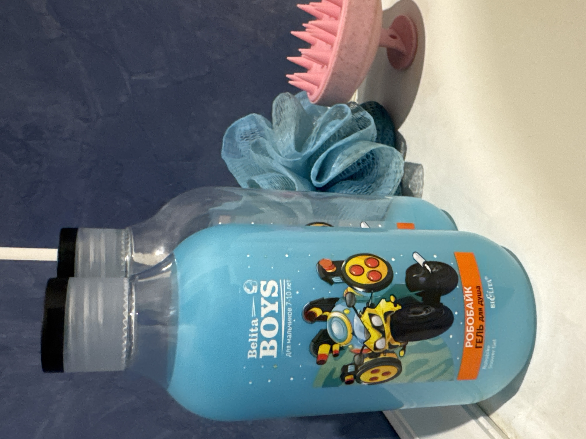 The image shows a close-up, eye-level, indoor shot of a blue liquid soap bottle with the brand name "Belita BOYS" and a cartoon robot motorcycle on it. The bottle is transparent, with blue liquid inside. To the right of the soap bottle, there is a blue mesh bath sponge and a pink scalp massager. In the background, a blue tiled wall is visible. The scene is set on a white surface, possibly a bathroom counter or shelf. The location is identified as Яромінскі сельскі Савет, Belarus, though there is no visual information in the image to confirm this specific location.