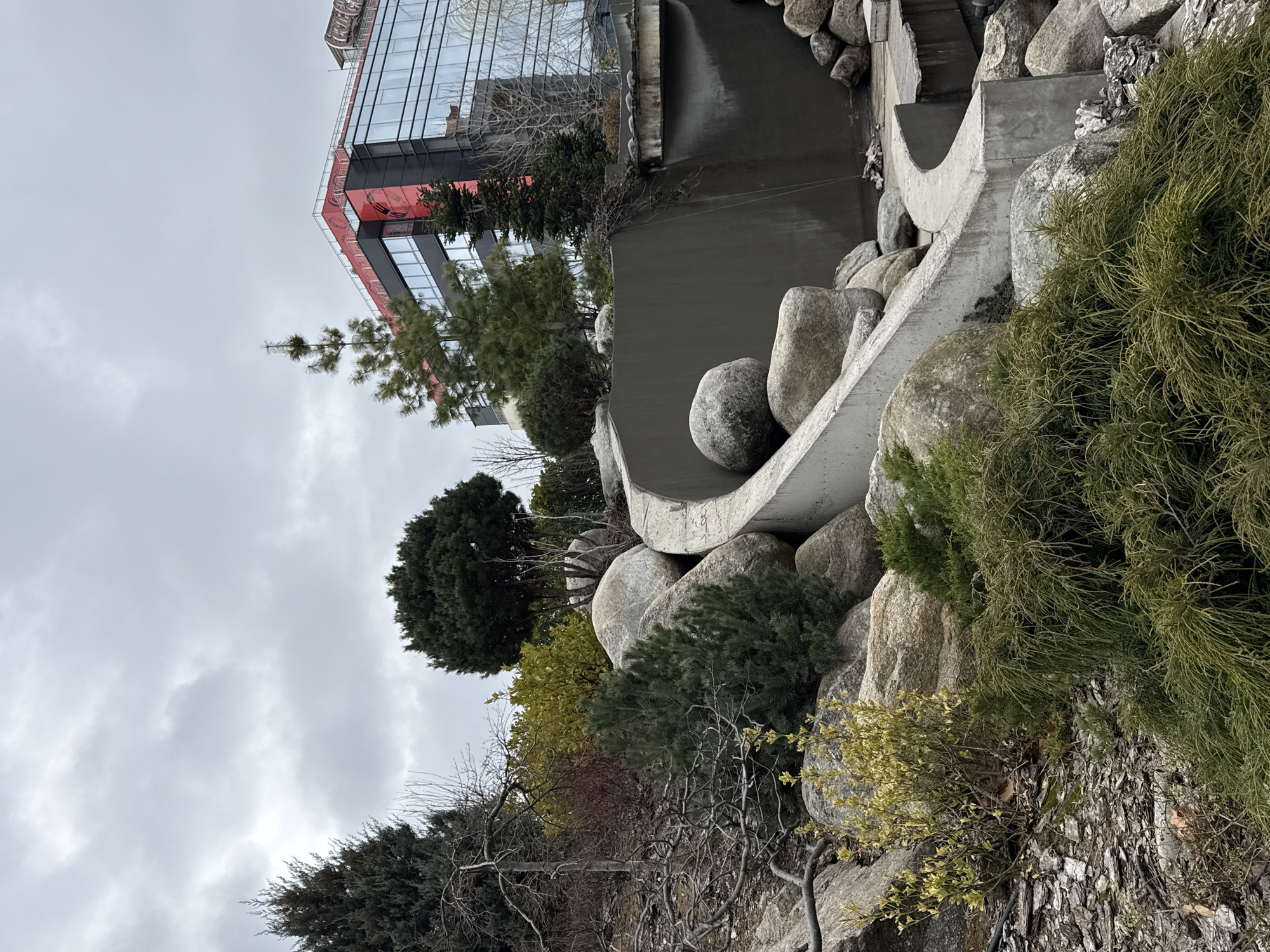 The image shows an outdoor scene with a combination of natural and artificial elements, likely an urban garden in Sofia, Bulgaria. In the foreground, there's a rock garden with various types of greenery including small shrubs, bushes, and a young tree. Larger rounded boulders are interspersed among the plants and a concrete structure featuring curved walls and channels. Water may be designed to flow through this structure. In the background, a modern building with glass windows and a red accent is visible, suggesting an urban setting. The sky is overcast. The photo appears to be taken from a low angle, possibly at ground level.