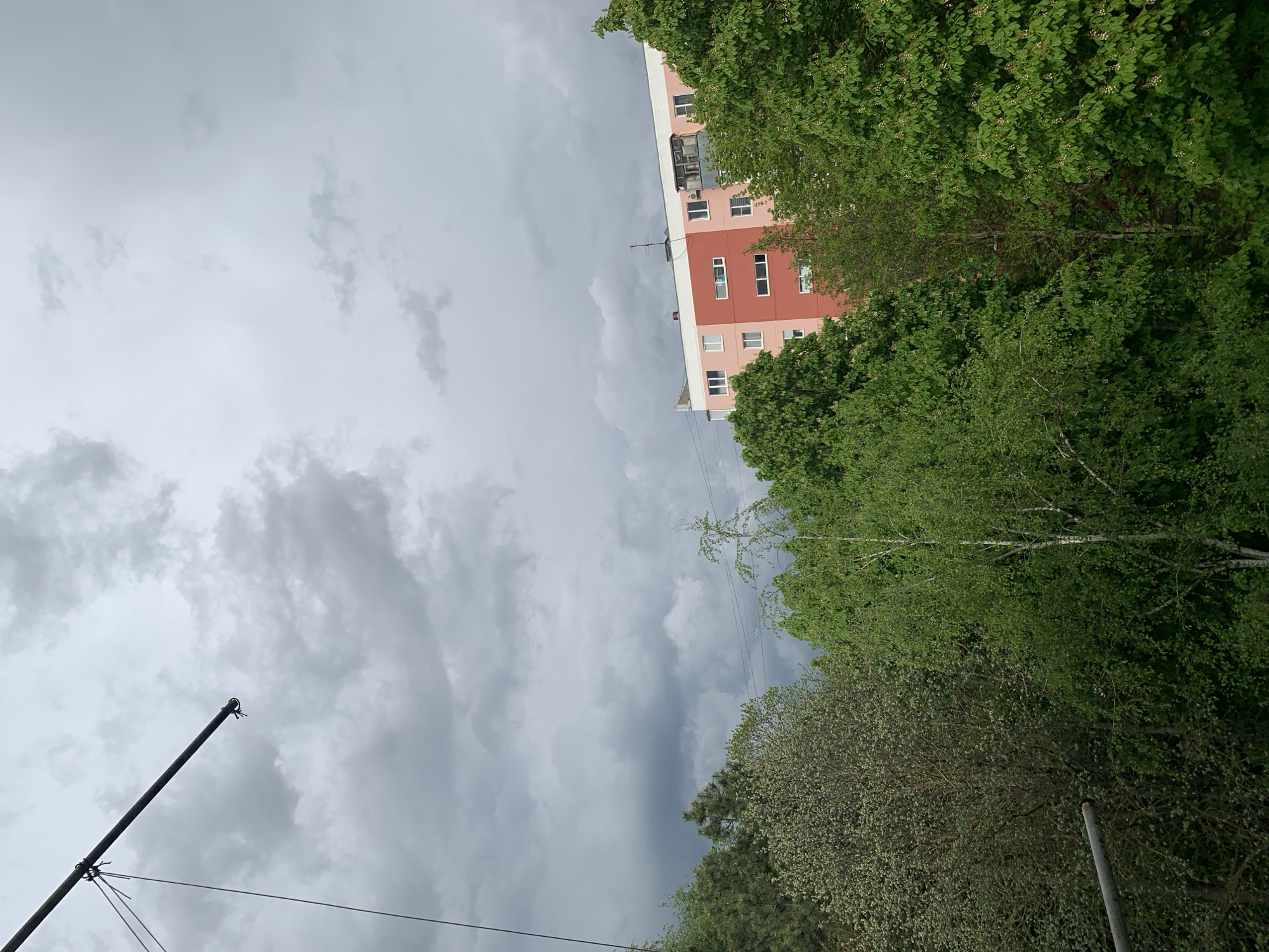 The image shows an upward-facing view of a cloudy sky and a building in an urban environment. The sky is dominated by gray clouds, suggesting an overcast or stormy condition. A red and pink building is partially visible, framed by green trees. A gray pole with wires attached is located in the lower left corner. Another gray pole stands in the lower right corner. The location appears to be in a city, though the specific name is unknown.