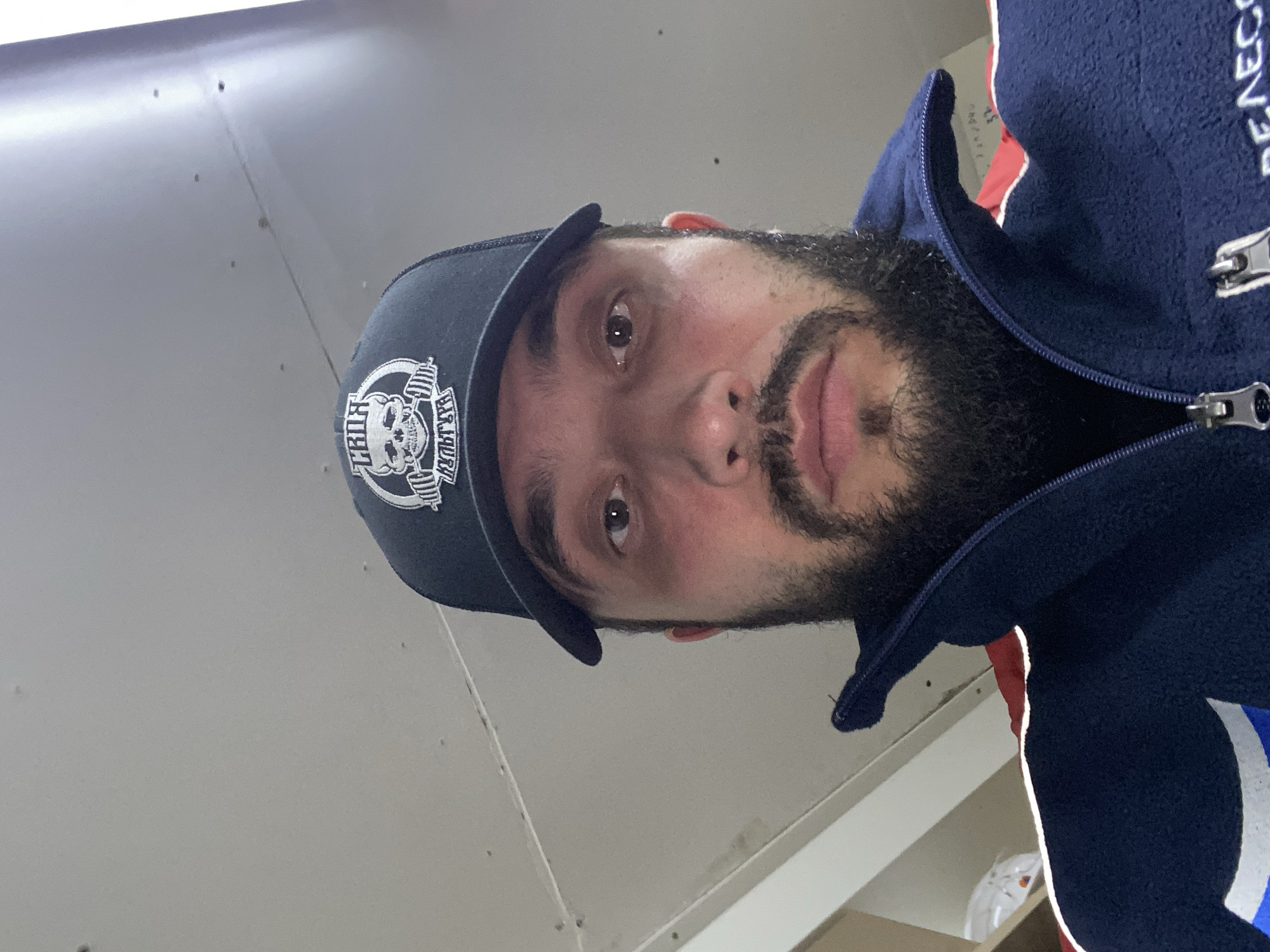 A selfie of a man wearing a black baseball cap with a skull logo and a dark blue jacket is taken from a low angle. The man has a beard and appears to be indoors, with a light-colored ceiling visible in the background. The image does not provide any specific information about the location within Luzhitsy, Russia, or any details that would suggest it is in Luzhitsy, Russia, beyond the prompt's assertion.