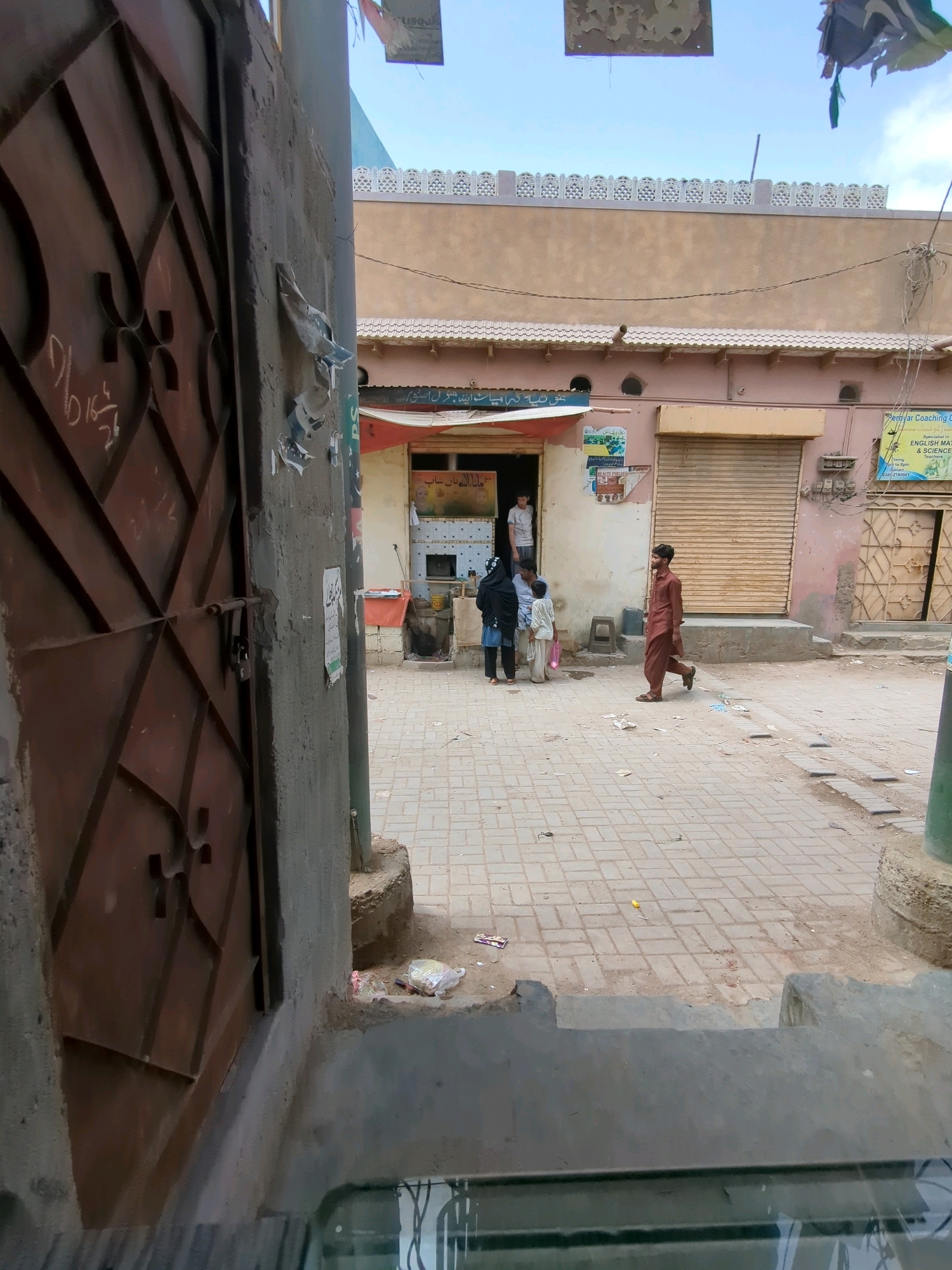 This is a street-level, eye-level, medium shot of a narrow alleyway in a densely populated urban area, possibly in Karachi Division, Pakistan. The time of day appears to be daytime, with bright sunlight casting shadows. The scene is characterized by its gritty, lived-in aesthetic.
In the foreground, to the left, is a large, dark brown metal gate with a patterned design and visible rust. It is padlocked. To the right of the gate is a textured, grey concrete wall with peeling posters and graffiti. A green pole is visible on the far right edge of the frame, possibly a lamppost or utility pole.
The alleyway itself is paved with interlocking grey bricks, showing signs of wear and tear. Scattered across the pavement are various pieces of debris, including plastic wrappers and what appears to be a yellow plastic bag.
In the middle ground, there are several buildings. One building on the left has an open doorway, with a man in a light-colored t-shirt and dark pants standing inside. Two children, one in a light-colored outfit and the other in a dark headscarf and blue garment, are standing in front of the doorway, possibly interacting with the man. A small red stool is placed near the doorway.
Further down the alley, to the right, is a building with a closed, corrugated metal shutter. A man in a maroon shalwar kameez is walking away from the viewer towards the right. Another building to the right of that has a closed metal door with a decorative pattern.
The architecture suggests a typical South Asian urban environment, with buildings constructed from concrete and plaster, often painted in muted colors. The presence of overhead wires and the general state of the street indicate a well-inhabited but perhaps economically challenged area. The overall impression is one of everyday life unfolding in a busy, potentially congested part of the city.