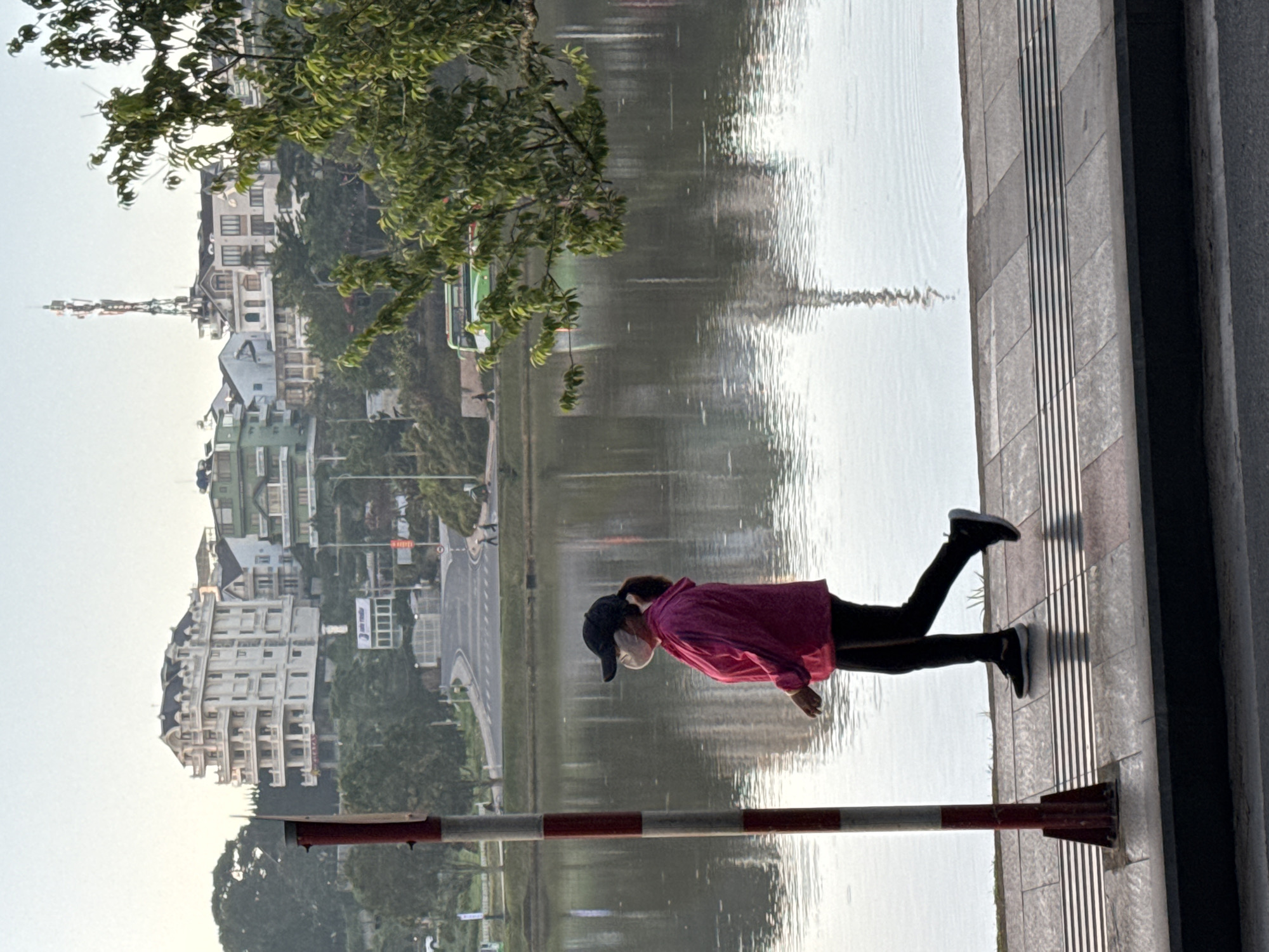 A woman wearing a pink hooded jacket, black pants, a black baseball cap, and a white mask is walking on a paved pathway next to a body of water. The woman is viewed from the side and is in motion. In the background, there are buildings and trees on a hillside. A green bus is visible on a road behind the water. The scene appears to be in Da Lat, Vietnam, possibly near Xuan Huong Lake, given the architectural style of the buildings and the presence of a large body of water within the city. The lighting suggests it is early morning or late afternoon.