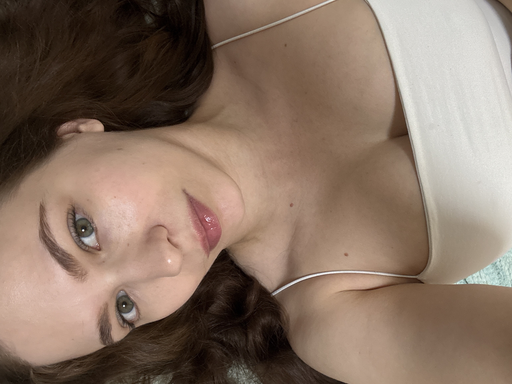 A close-up, high-angle shot captures a person, likely a young adult female, lying down. Her head is tilted, and she is looking directly at the viewer with serene green eyes. Her long, wavy dark brown hair is spread out around her head. Her eyebrows are well-defined, and her lips are adorned with a glossy, light pink product. She has clear skin with a few small moles visible on her neck and shoulder area.
She is wearing a light-colored, possibly beige or off-white, top with thin spaghetti straps, which exposes her shoulders and cleavage. The fabric of the top appears soft and slightly textured. Parts of her neck, collarbone, and upper chest are prominent in the frame. The background is largely obscured by her hair and body, with a subtle hint of a light blue-green textured surface beneath her. There is no visible text in the image. The activity appears to be posing for a photograph while lying down. The location context for this image is Chaikovsky, Russia.