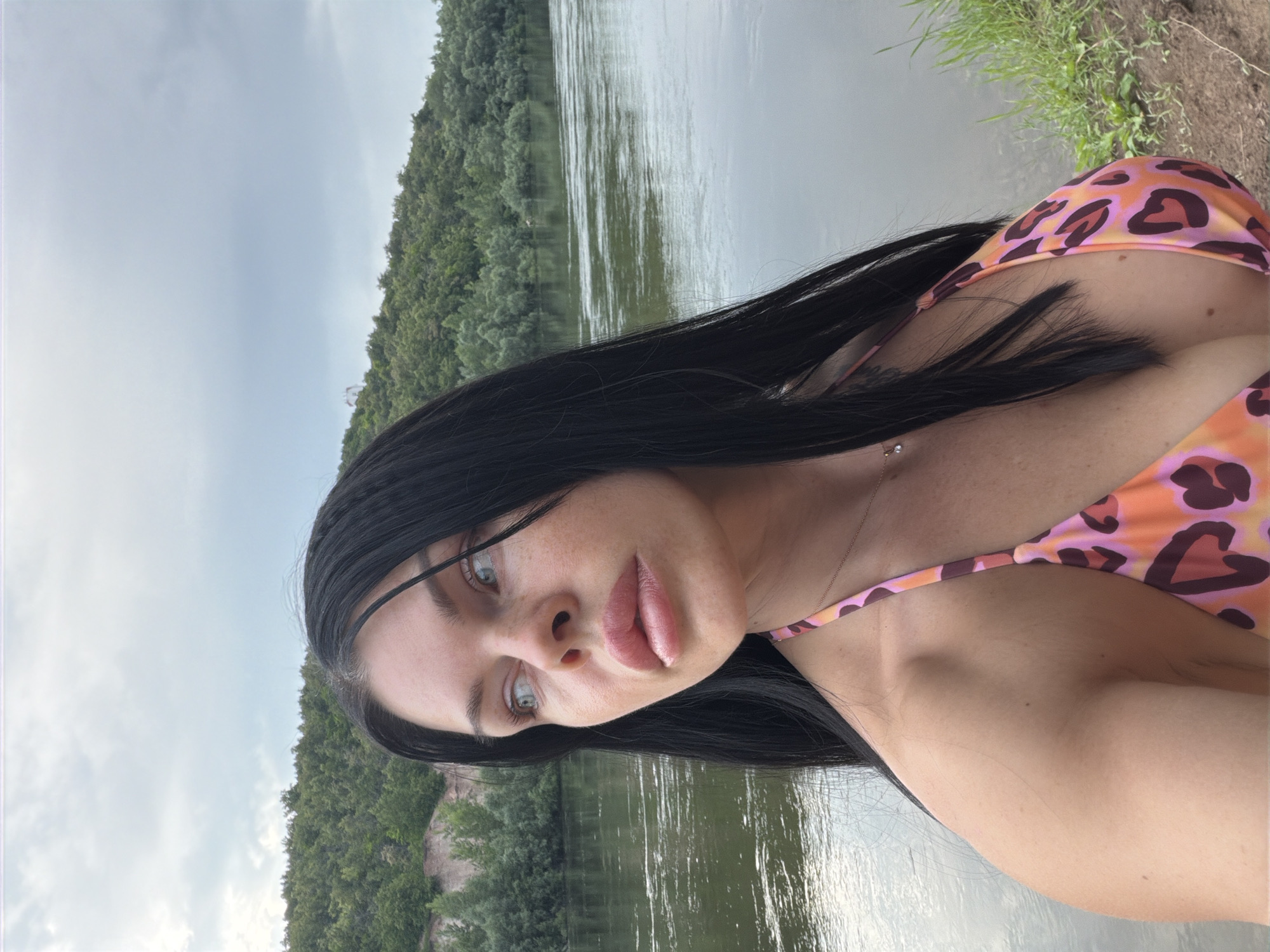 The media file is a portrait of a young woman with long, dark hair, taken outdoors near a body of water. She is wearing a leopard-print bikini in shades of orange and pink. The setting appears to be a natural landscape, with a river or lake in the background, bordered by a dense forest of green trees. The sky is overcast, suggesting a cloudy or potentially rainy day. The woman is looking towards the camera with a neutral expression. Her features are visible, including her eyes, nose, and lips. A delicate necklace with a small pendant is visible around her neck. The image has a vertical orientation. The context provided indicates the location is Ufa, Russia.