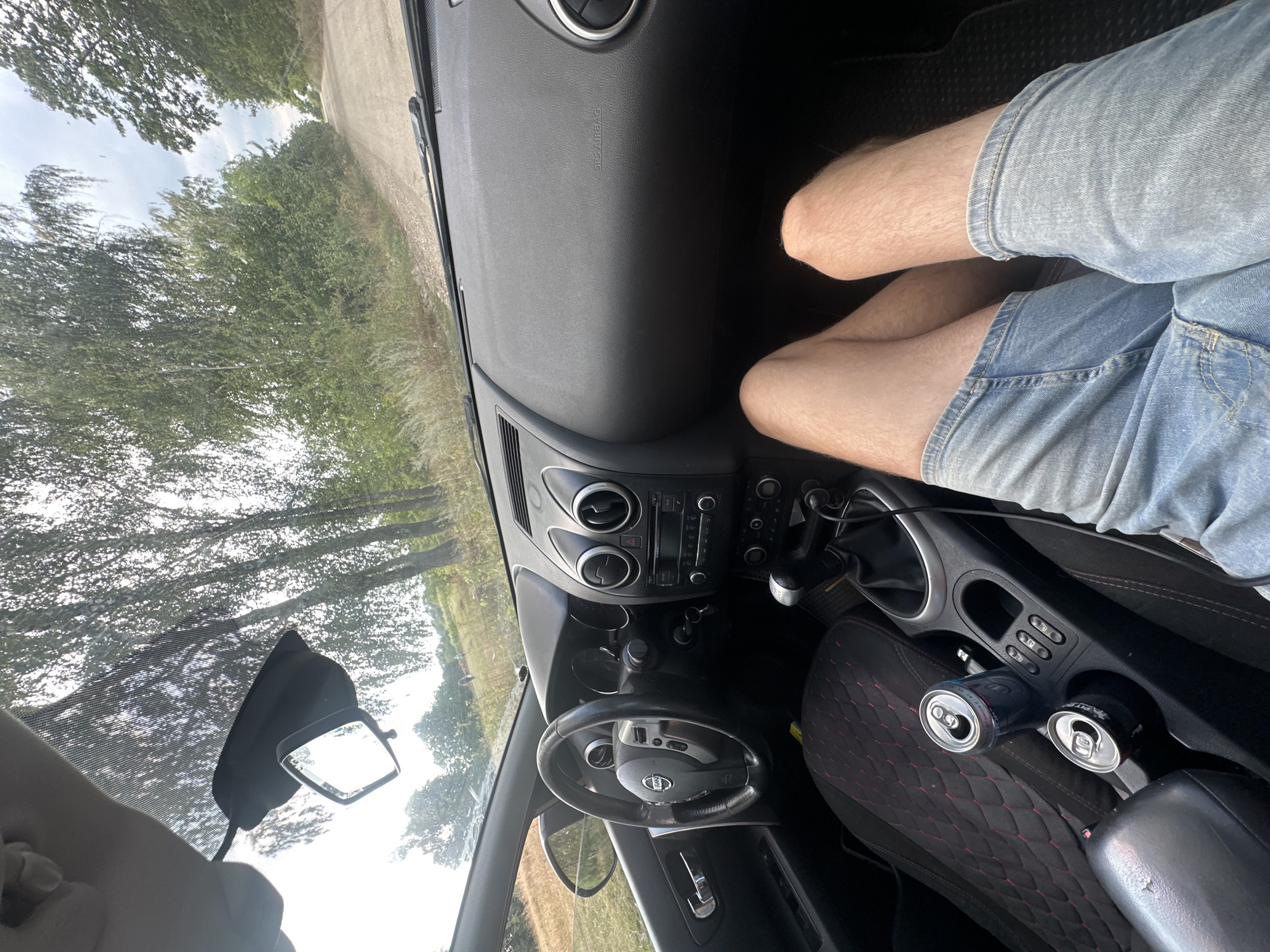 A person's legs are visible in the foreground, wearing denim shorts. They are seated in the driver's seat of a car, with the dashboard and steering wheel to the left. Two open cans are in the center console. The view through the windshield shows a dirt road surrounded by trees and vegetation, suggesting a rural or undeveloped area. The sky is overcast.