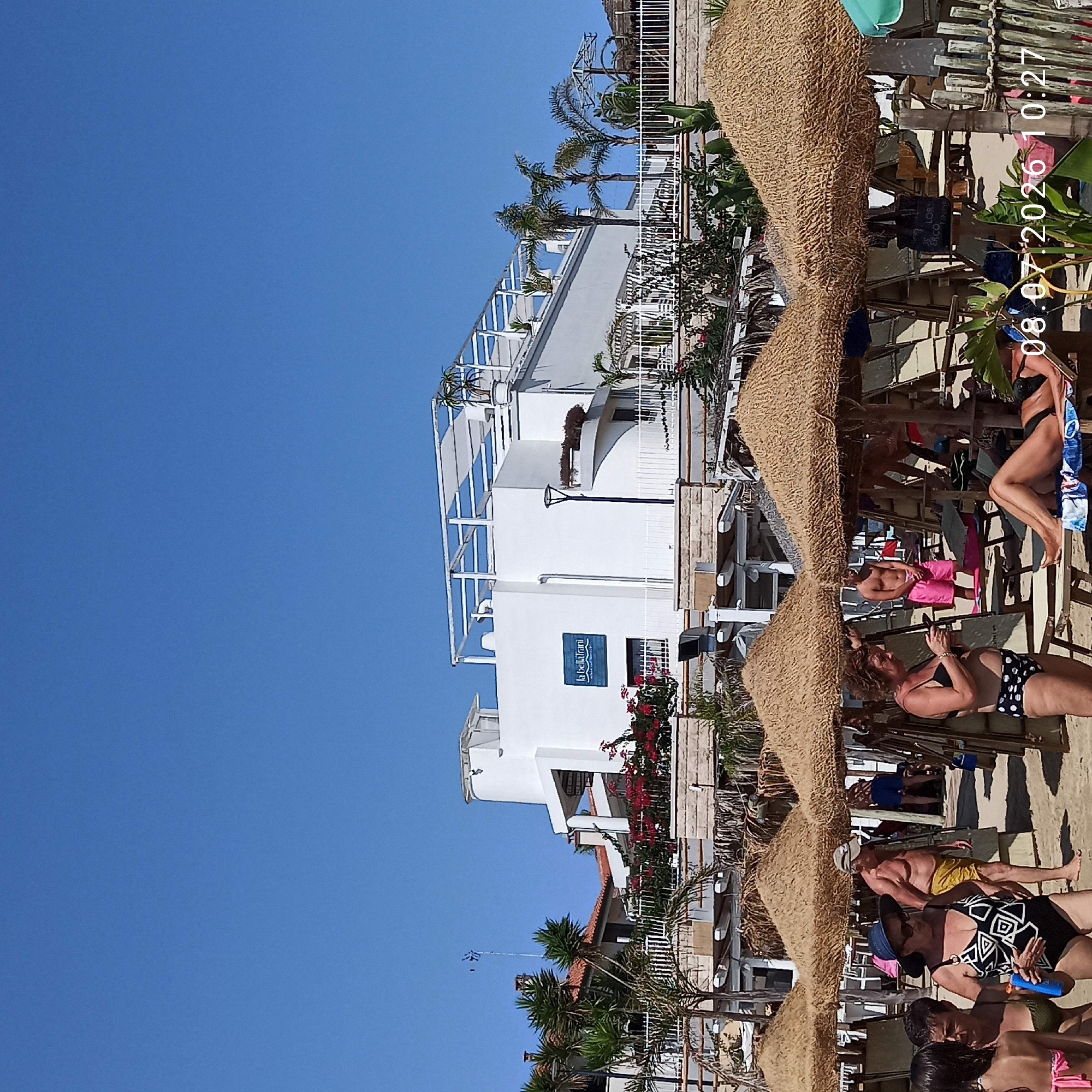 The image depicts a sunny day at a beach in Trani, Italy. In the foreground, several beach umbrellas with straw coverings are erected, providing shade. People are seen in various states of relaxation and activity. A woman in a black and white patterned swimsuit and a blue hat is standing, holding a blue bottle. Another woman in a black bikini with white polka dots is looking at her phone. A man in yellow shorts and a baseball cap is walking. In the background, a white building with a blue sign that reads "la Bellarani" is visible, likely a hotel or resort. Palm trees and lush green plants surround the building, adding to the tropical atmosphere of the beach. The sky is a clear, bright blue. The timestamp "08.07.2026 10:27" is superimposed on the lower right corner of the image.