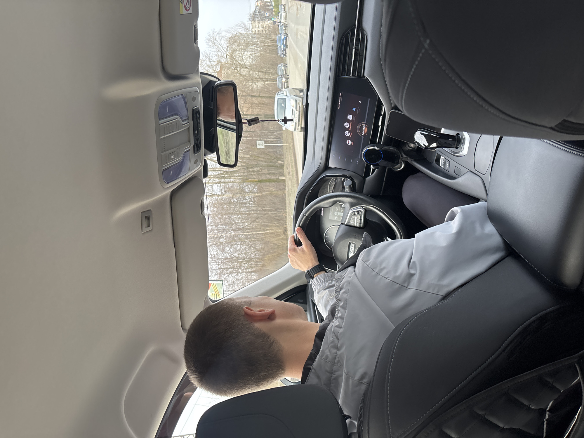 A Caucasian male with short brown hair is driving a grey Haval Jolion car. He is wearing a grey jacket and a black wrist watch. The car is on a road with other vehicles in front, including a white van. There are trees and buildings in the background. The interior of the car features a touchscreen display with the Haval logo, a rearview mirror with a cross hanging from it, and grey leather seats. The location is indicated as Chaikovsky, Russia, based on possible context clues of make and model availability and external surroundings typical of Russian urban areas.