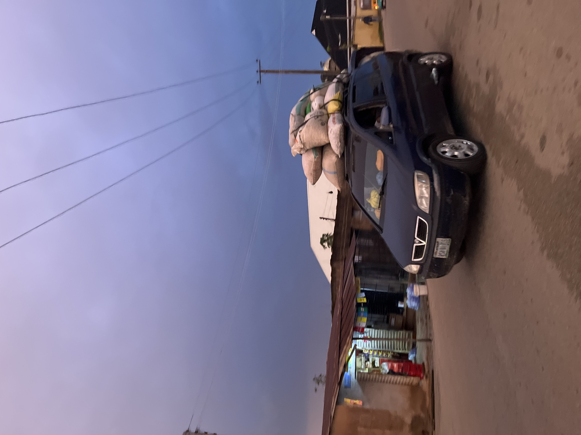 The image depicts a street scene at dusk in the city of Jos, Nigeria. A dark blue minivan is the central subject, traveling on a paved road. The vehicle's roof is heavily laden with a large stack of sacks, secured with ropes. The sacks appear to be filled with grain or similar dry goods, with varying shades of beige and white. The minivan has its headlights on, illuminating the road ahead. Inside the vehicle, a person is visible in the driver's seat.
To the left of the road is a row of small market stalls with corrugated metal roofs. One stall is brightly lit from within, showing shelves stocked with items, and a red cylindrical object, possibly a fire extinguisher or a large bottle, is visible outside. Another stall appears to be more open, with items displayed. Various plastic containers, some filled with water, are placed near the stalls.
In the background, the sky is a deep twilight blue, with thin clouds. Several utility poles with power lines crisscross the sky, casting long shadows. Further in the background, there are buildings, including one with a white, angular roof. A small, yellow auto-rickshaw or similar vehicle is seen at the far right of the frame, with a person walking nearby. The road surface is wet in patches, suggesting recent rain. The overall impression is a busy, everyday scene in a Nigerian urban environment during the evening hours.