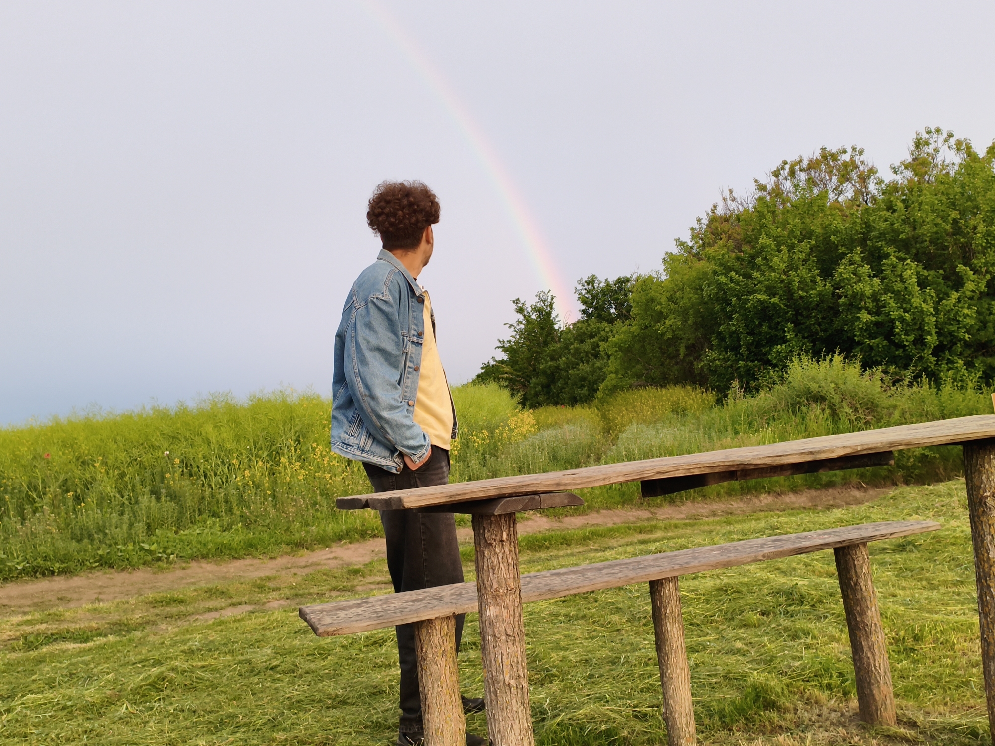 A person stands near a wooden table and bench structure, looking towards a rainbow in a rural setting. The person has dark curly hair and is wearing a yellow shirt with a denim jacket and dark pants. To the left is a field of green and yellow vegetation, and to the right, a forest of green trees. The background sky is light. The image was taken in Ustyvytsia, Ukraine.
