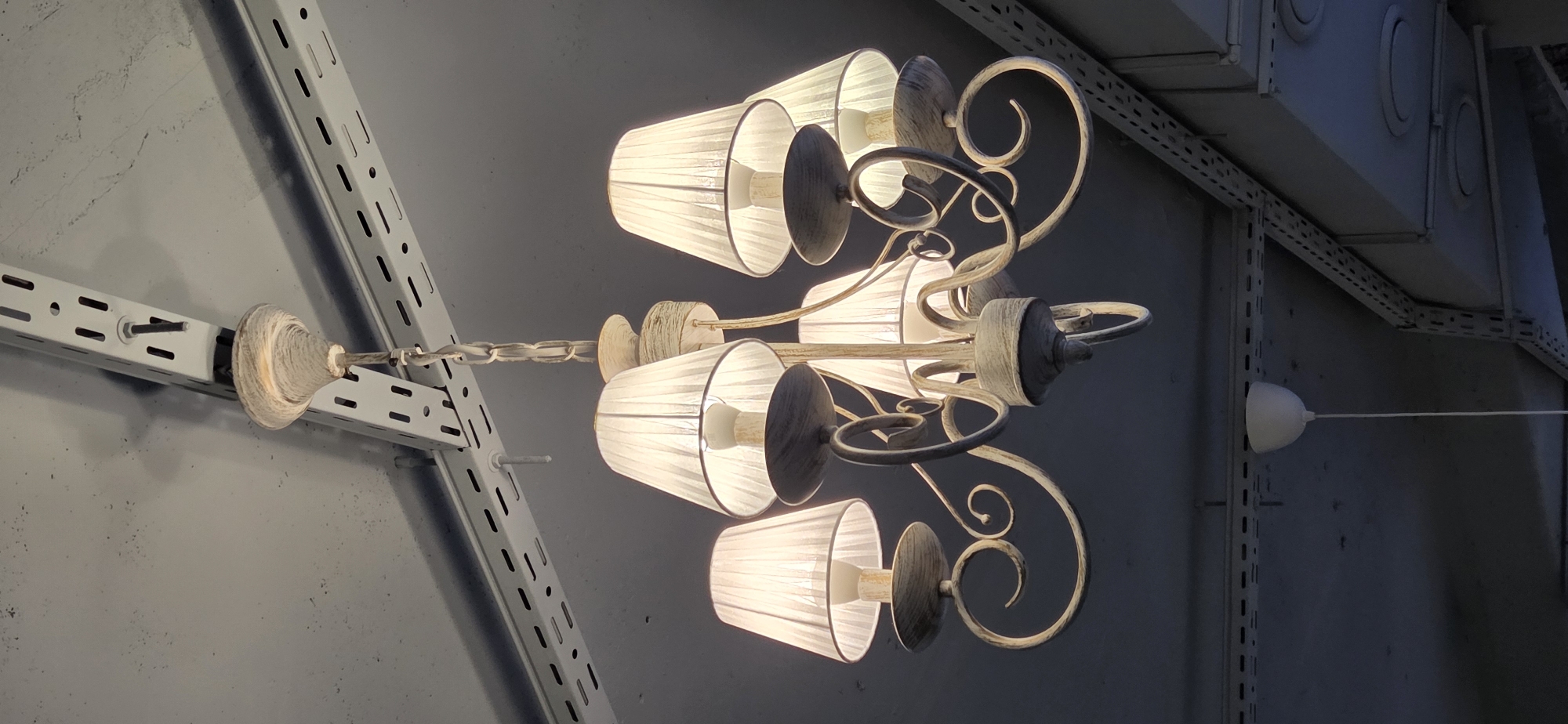 An upward-angled view captures a six-light chandelier suspended from a ceiling in Lipetsk, Russia. The chandelier features a distressed, off-white painted metal frame with ornate, scrolling arms. Each arm supports a vertically pleated, translucent white fabric shade, revealing an illuminated incandescent bulb within. The chandelier hangs from a chain connected to a decorative, bell-shaped ceiling cup, also in a distressed finish. The ceiling is a light gray or off-white surface, showing minor discoloration and texture. Running across the ceiling and extending into the background are multiple parallel runs of white, perforated metal channel struts, characteristic of industrial shelving or mounting systems, secured with visible bolts. To the lower left of the main chandelier, a smaller, plain white bell-shaped ceiling rose or fixture cover with a thin white cord hangs down. In the background on the right, more structural elements and circular fixtures are partially visible. The overall lighting is dim, highlighting the illuminated chandelier.