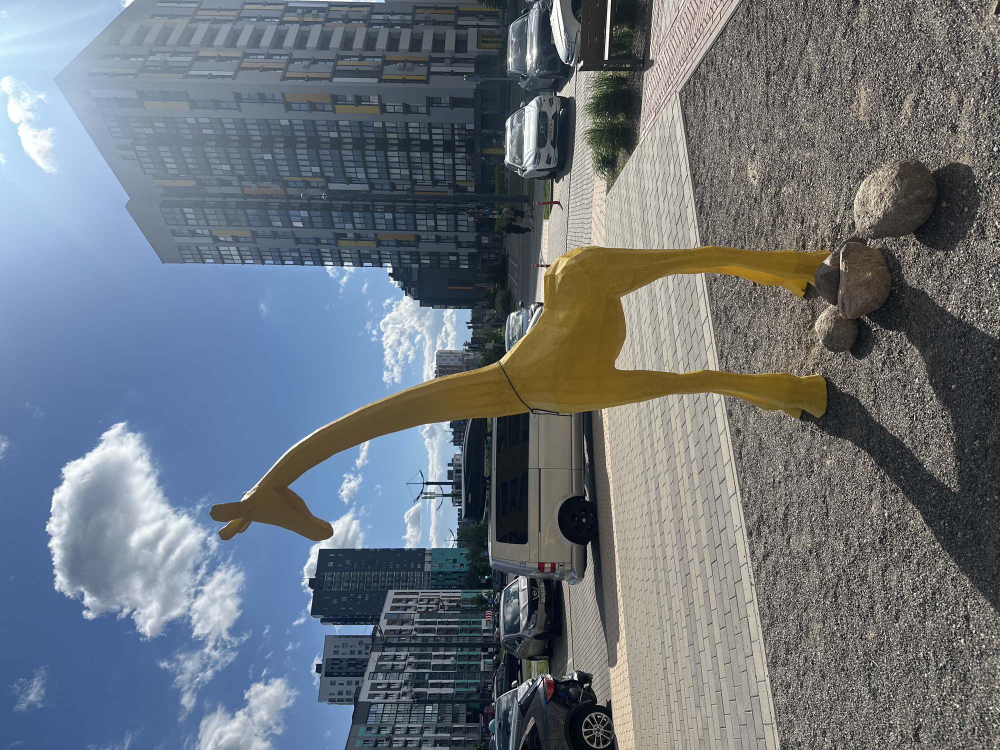 A yellow giraffe statue lies horizontally on a stone sidewalk. Three rocks rest on the ground to the right of the giraffe's legs. The city of Бараўлянскі сельскі Савет, Belarus, is visible in the background, with tall apartment buildings, cars, and a blue sky with scattered clouds. A light-colored van and several cars are parked behind the giraffe.