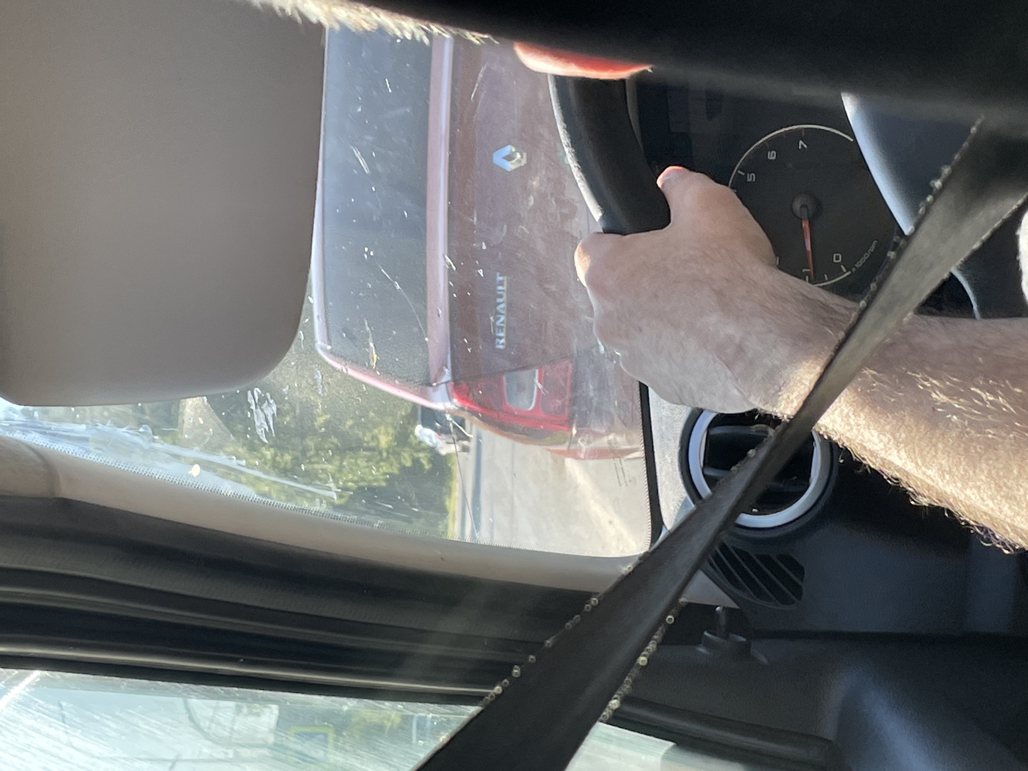 The image shows the interior of a car, likely a Renault, as indicated by the logo and text on the vehicle in front. The perspective is from the driver's seat, with a hand on the steering wheel and part of the dashboard visible. The driver is wearing a seatbelt. Outside the car, traffic appears to be moving slowly, with another car in front. The scene suggests a typical day in a city, possibly Chaikovsky, Russia, given the context. The lighting indicates it is daytime.