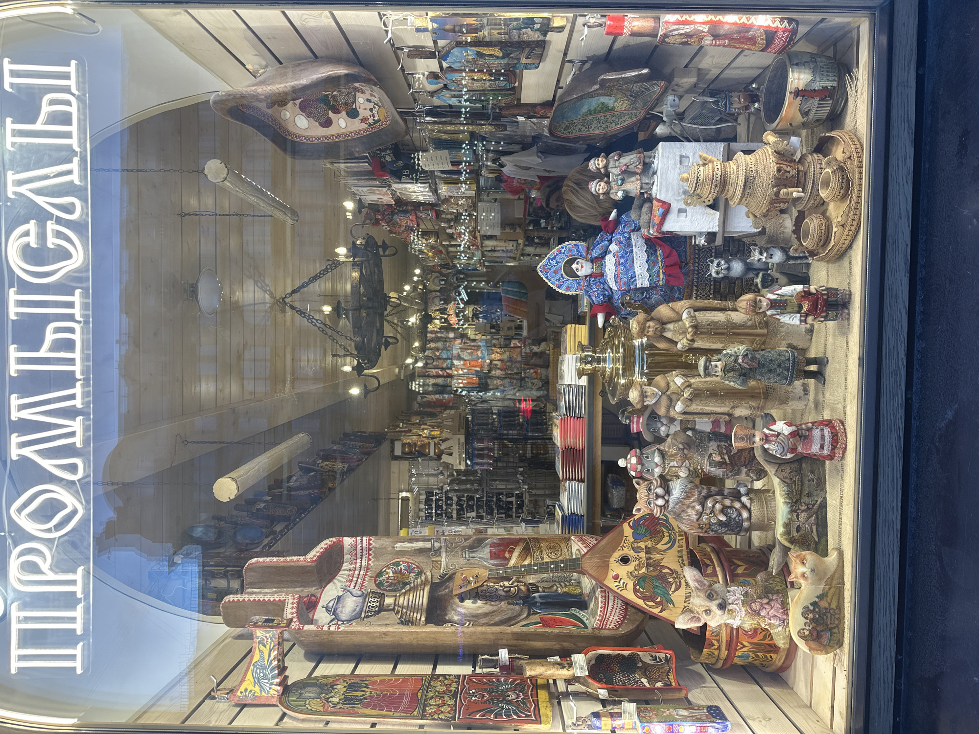 The image depicts a storefront window display, likely in Saint Petersburg, Russia, judging by the merchandise. The window is filled with traditional Russian crafts and souvenirs, including wooden bowls, carved figures, samovars (tea urns), dolls, and lacquer boxes. There are various items densely packed in the display. The storefront has a white neon sign with Cyrillic letters on the left side of the window. The interior of the shop is visible through the window, showing white paneled walls and a chandelier. The display is arranged on wooden shelves. Reflections on the window surface show external elements like a building and possibly other street features, suggesting the shop is located on a street.