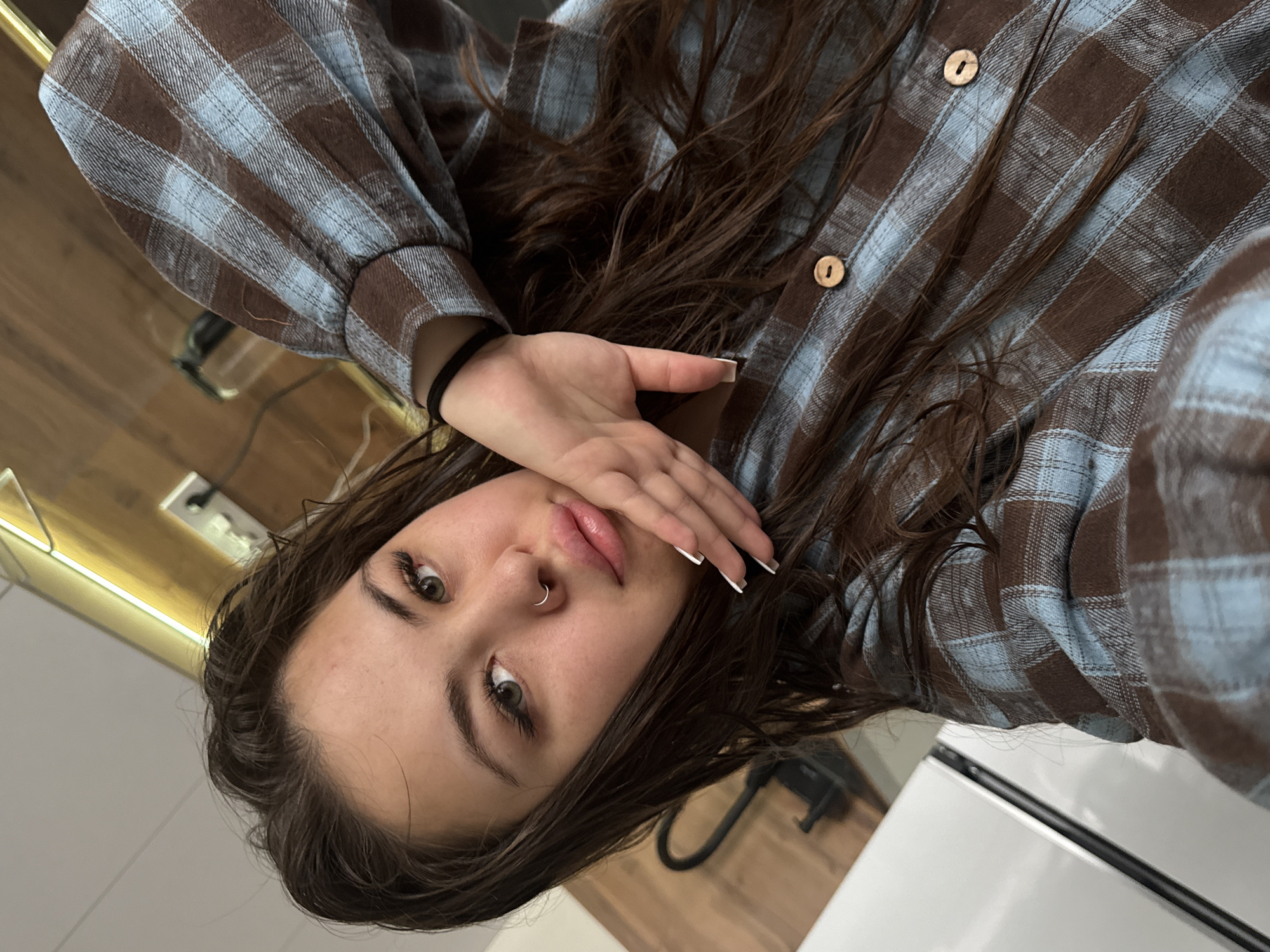 A rotated close-up portrait captures a young woman looking directly at the camera, positioned indoors. She is seen wearing a long-sleeved plaid shirt featuring a pattern of brown and light blue squares. Her long, dark brown hair, appearing slightly damp or styled with a casual texture, is scattered around her shoulders and against the shirt. Her right hand is raised, partially obscuring the lower part of her face, revealing manicured nails with white tips. She sports a silver hoop nose ring and has a black elastic hair tie on her left wrist. The background elements suggest a contemporary interior setting, possibly a kitchen or living area, with warm-toned wooden panels, a horizontal strip of artificial lighting on the left, and clean white surfaces. The subject's expression is subtle and direct. No text is visible in the image. The scene offers a personal glimpse, set in Bugry, Russia.