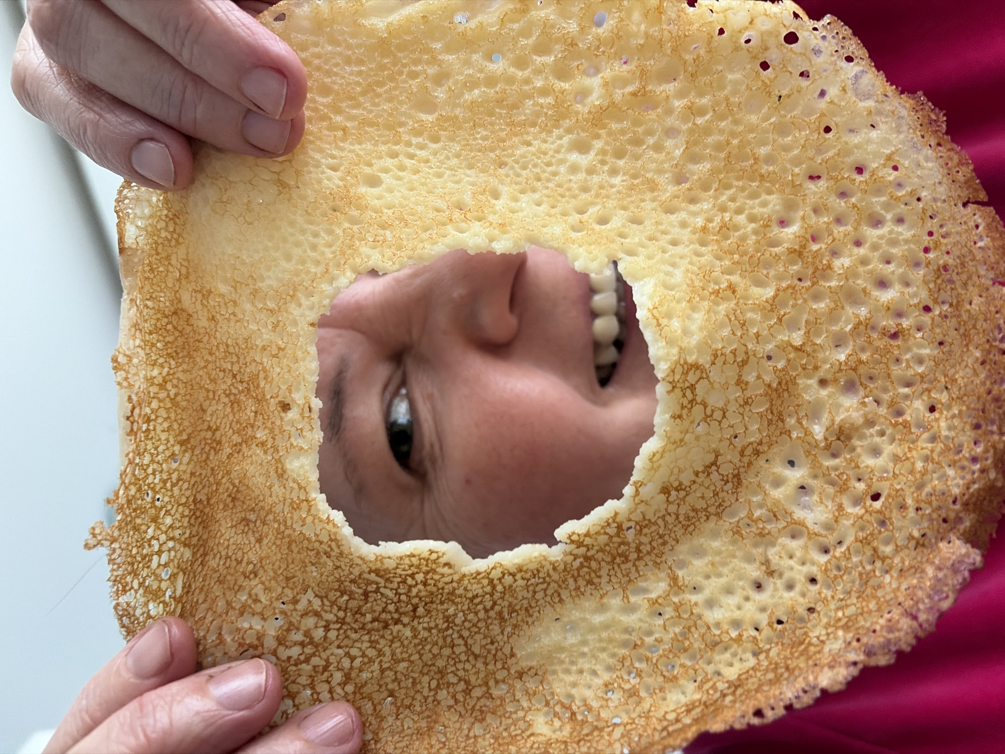 A person's face peers through a large, porous, circular crepe. The crepe is held up by two hands with manicured nails. The person's face is upside down. They are smiling, revealing their teeth. The background is a mix of off-white and a dark pink fabric, possibly clothing. No information indicates this photo was taken in Neftekamsk, Russia.