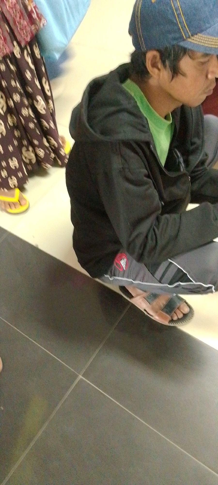 The image shows a person from the waist down wearing a black hooded jacket, a green t-shirt, grey track pants with white stripes, and brown and black sandals. The person is seated on a light-colored tiled floor, with dark grey tiled flooring in the foreground. To the left of the person, another individual is visible, wearing a patterned dress with brown and lighter colored floral designs and yellow flip-flops. The background appears to be an indoor setting with a light blue fabric draped in the upper left corner. The scene is consistent with an interior location in Sumenep, Indonesia, possibly a waiting area or a public space.
