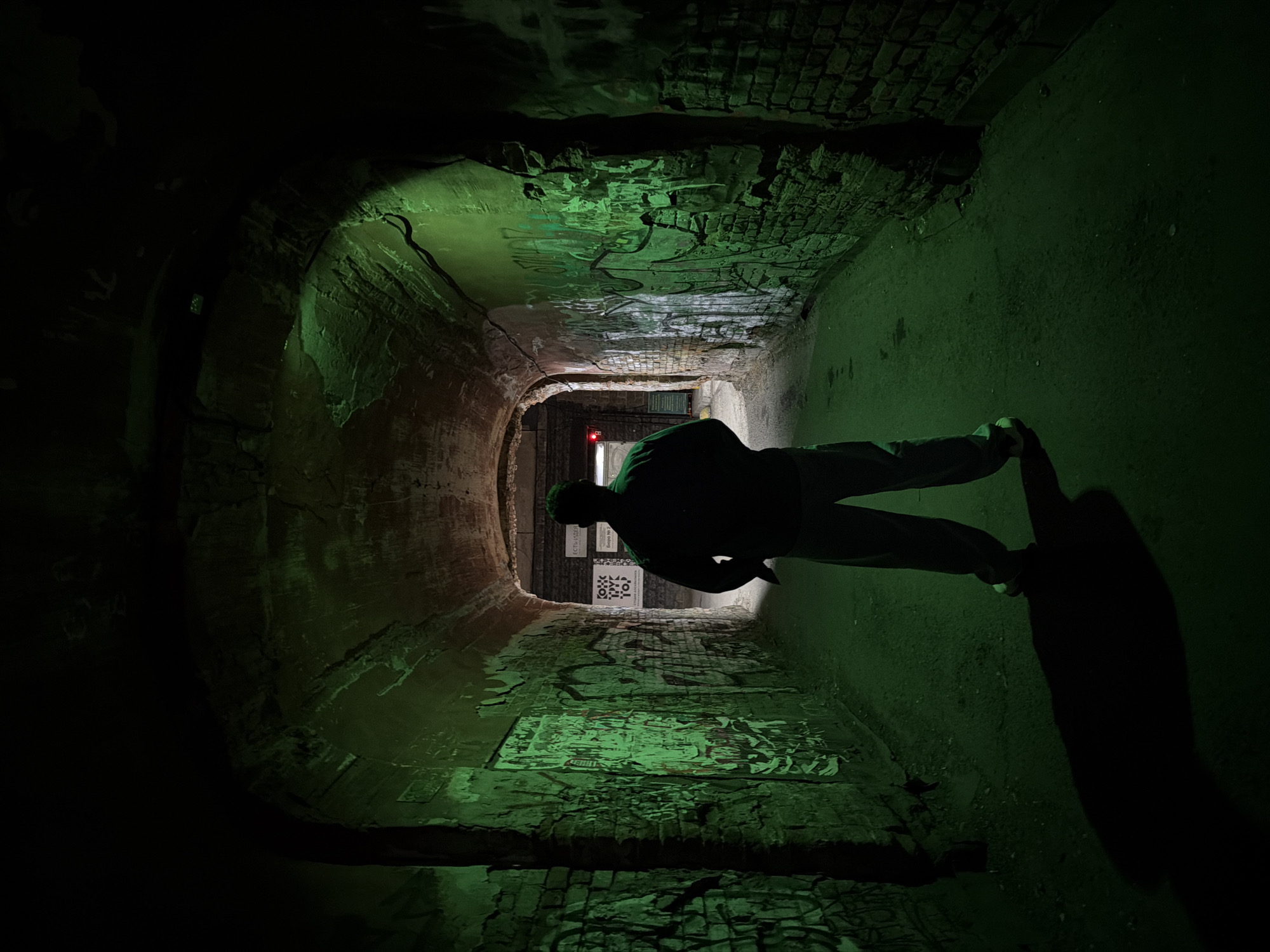 A dimly lit, arched brick tunnel with graffiti covers the walls. A figure stands within the tunnel, their form largely silhouetted and casting a long shadow. At the far end of the tunnel, a sign and a red light are visible. The lighting in the tunnel is predominantly green. This is the pedestrian tunnel near ulitsa Karla Marksa, 64 in Krasnoyarsk, Russia.