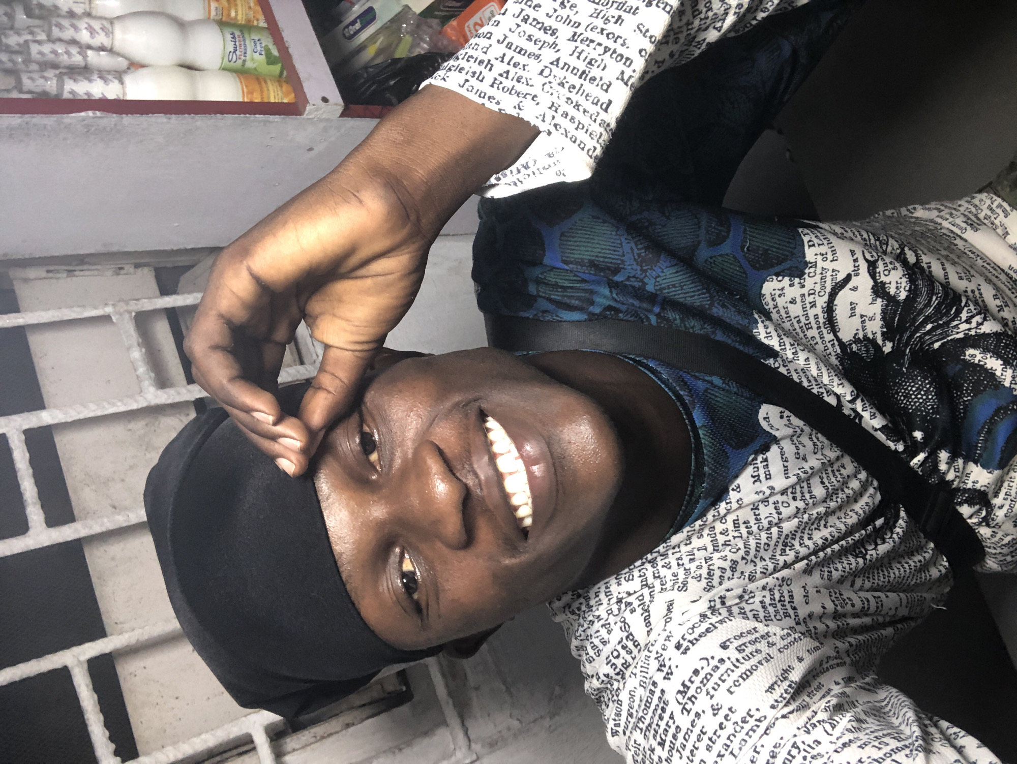 A selfie taken by a dark-skinned young man wearing a black head covering and a white shirt with text printed on it, stands in front of a white barred window. He is smiling broadly and has his right hand on his forehead. In the background, on the right side of the image, a shelf with several bottles of what appears to be juice or milk is visible. The setting is likely a commercial establishment in Ado, Nigeria, possibly a small shop or market stall. The lighting suggests an indoor environment with some artificial light source.